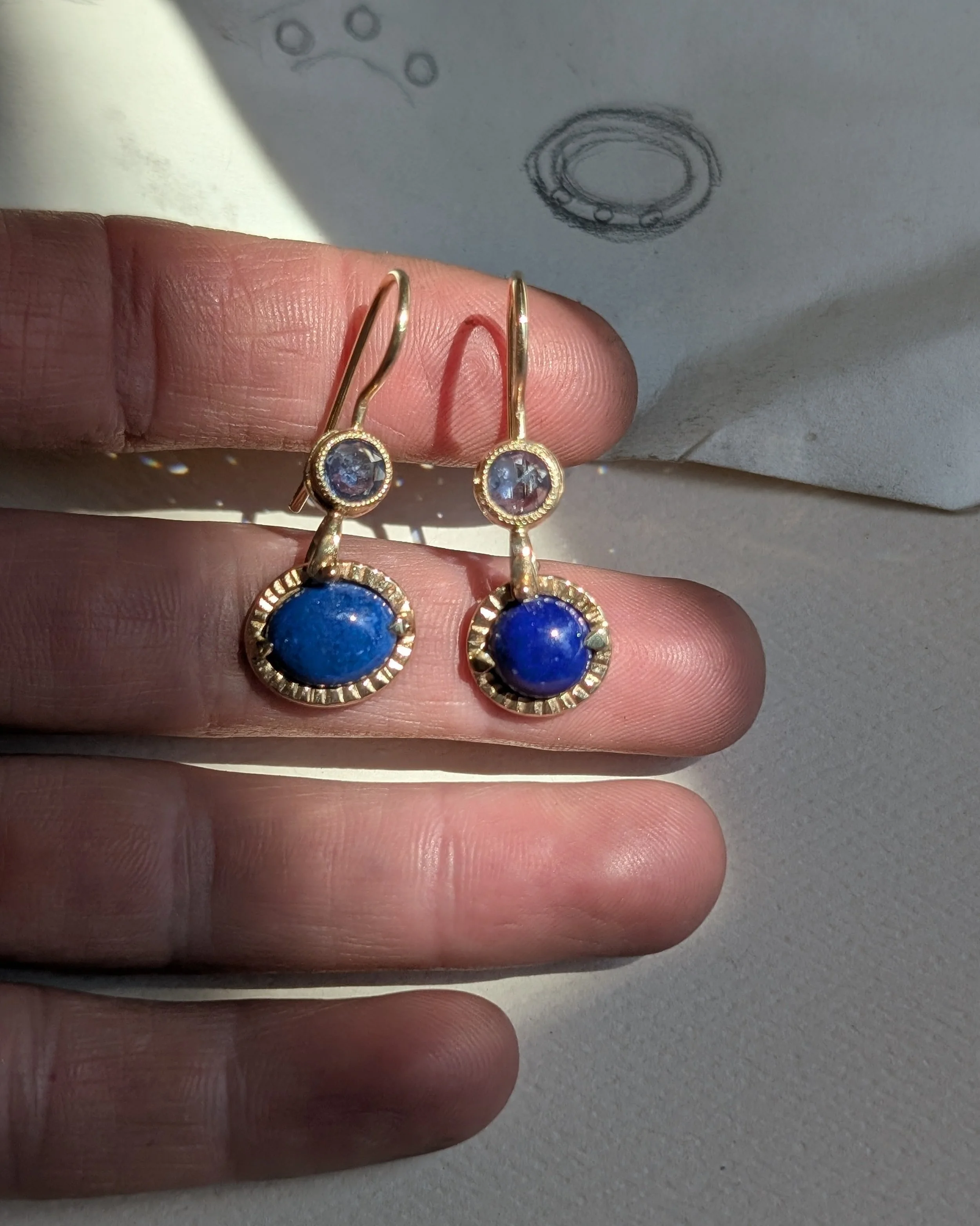 A pair of gold drop earrings with a small round gemstone at the top and a larger blue oval gemstone at the bottom, held against a person's hand with a sketch of a jewelry design in the background.