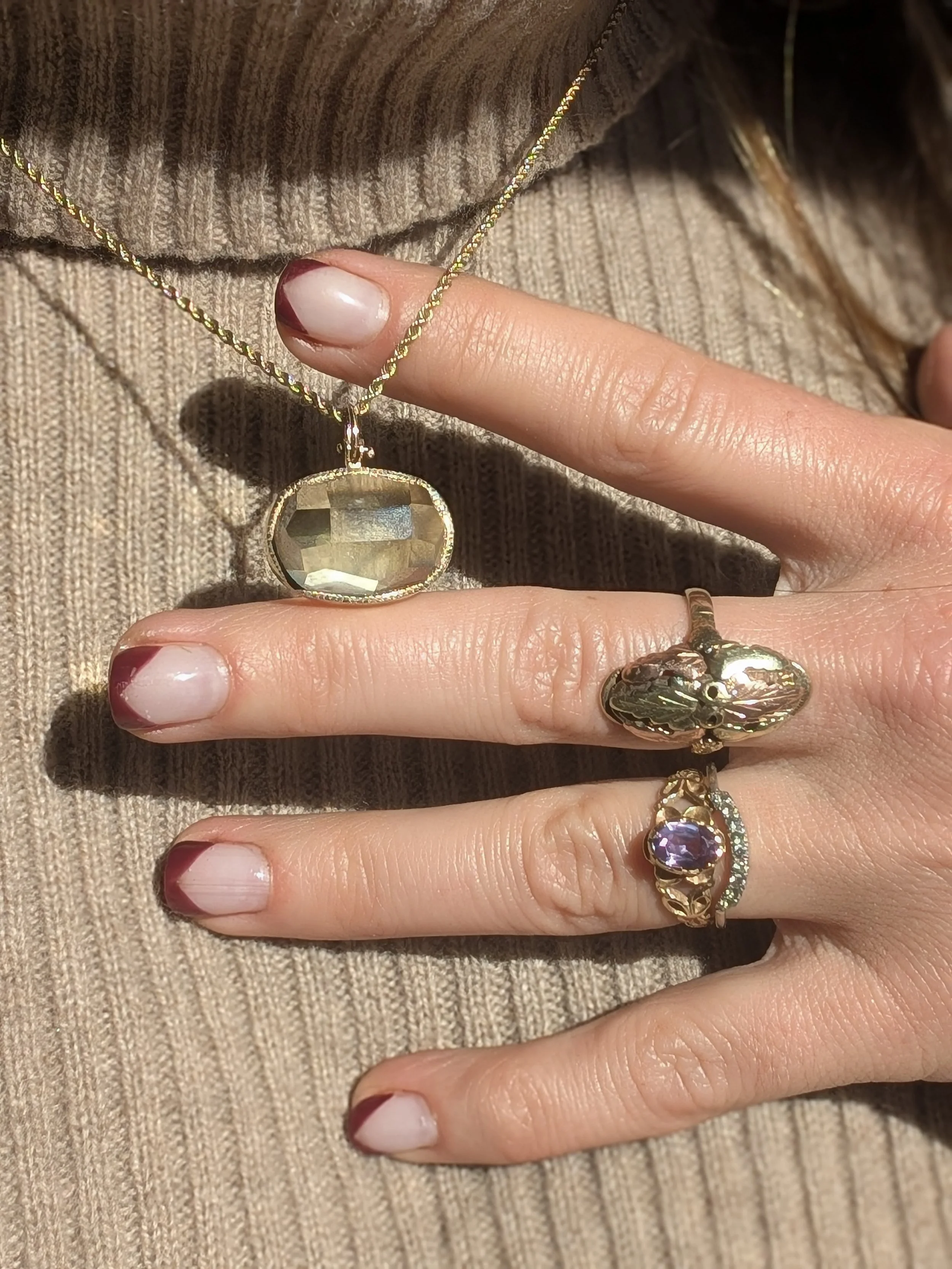 Close-up of a hand wearing multiple rings, a necklace, and a beige ribbed sweater. The hand has painted fingernails with a French tip design.