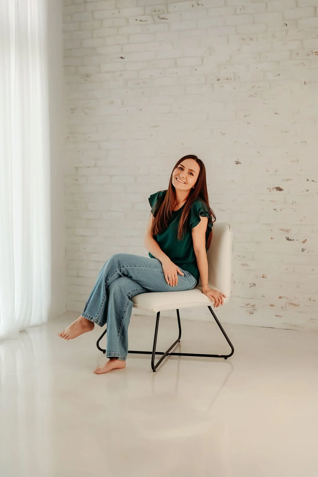 A woman sitting on a white chair in a bright room with a white brick wall and white curtains, smiling and barefoot.