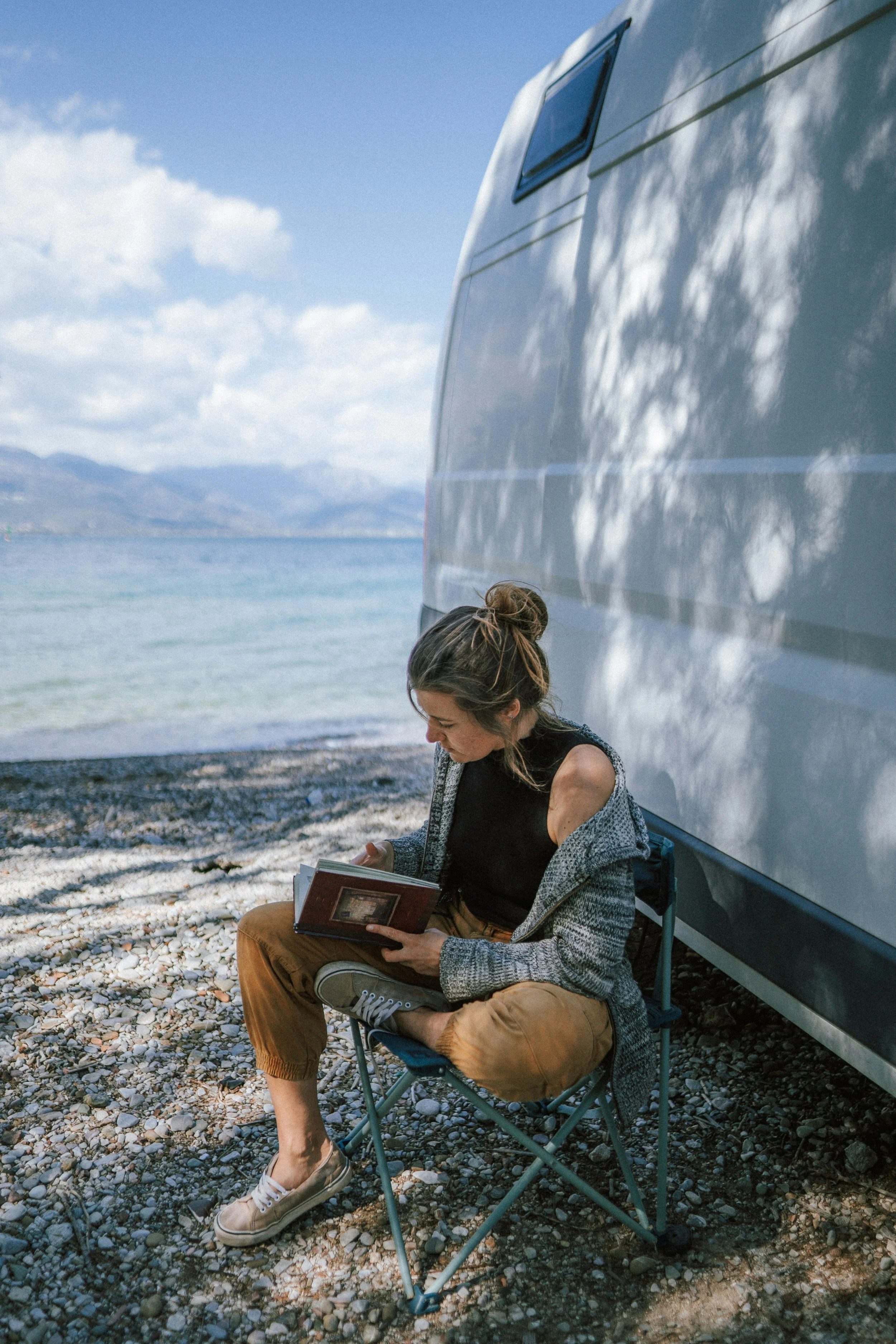 A young woman sitting on a camping chair beside a camper trailer, reading a book near a lake under a partly cloudy sky.
