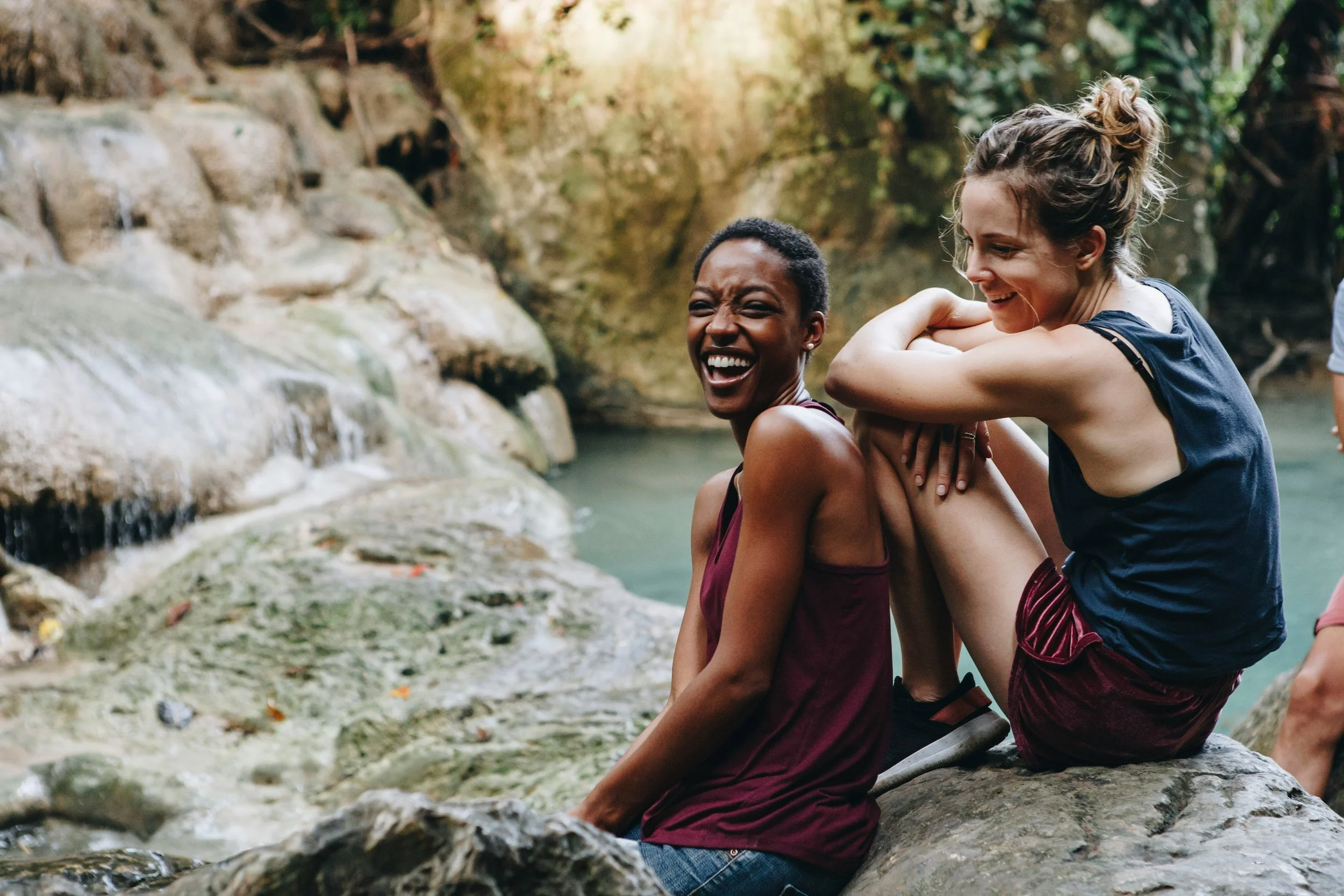 Two women sitting on rocks by a waterfall, smiling and enjoying each other's company in a natural outdoor setting.