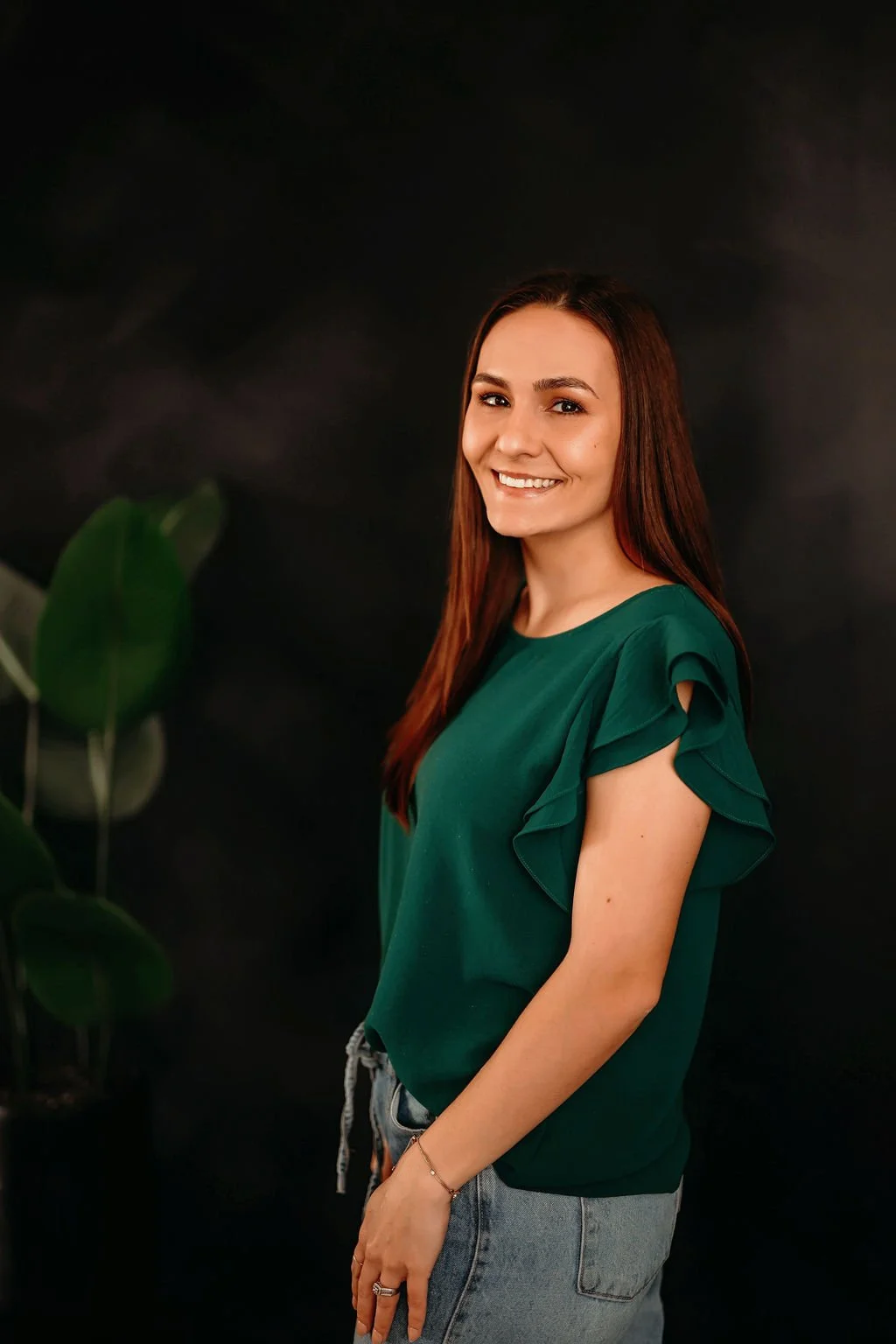 A young woman with long brown hair wearing a green blouse and jeans, smiling at the camera, standing in front of a dark background with green leaves on the left side.