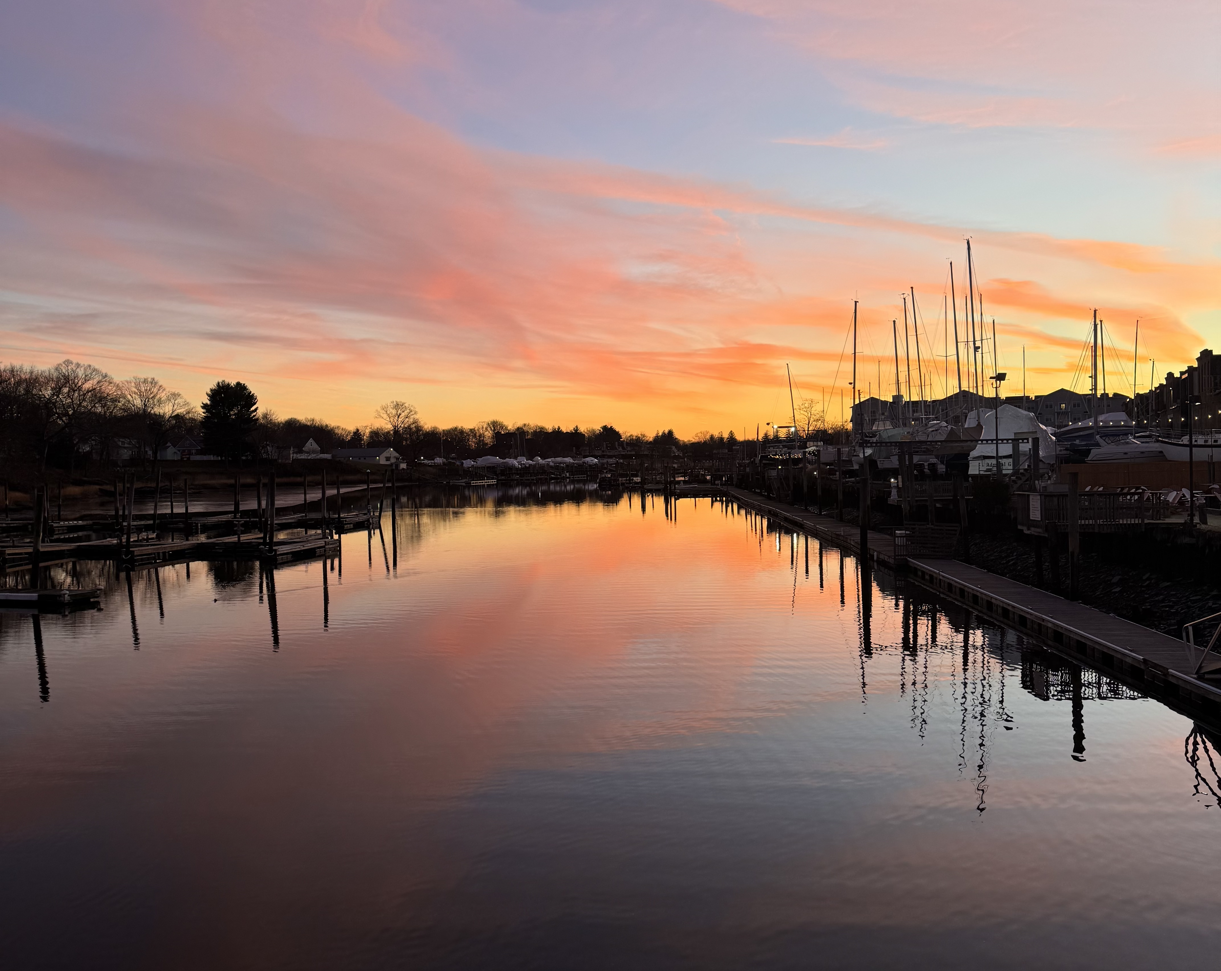 Sunset over a marina with calm water reflecting the colorful sky and boats docked along the pier.