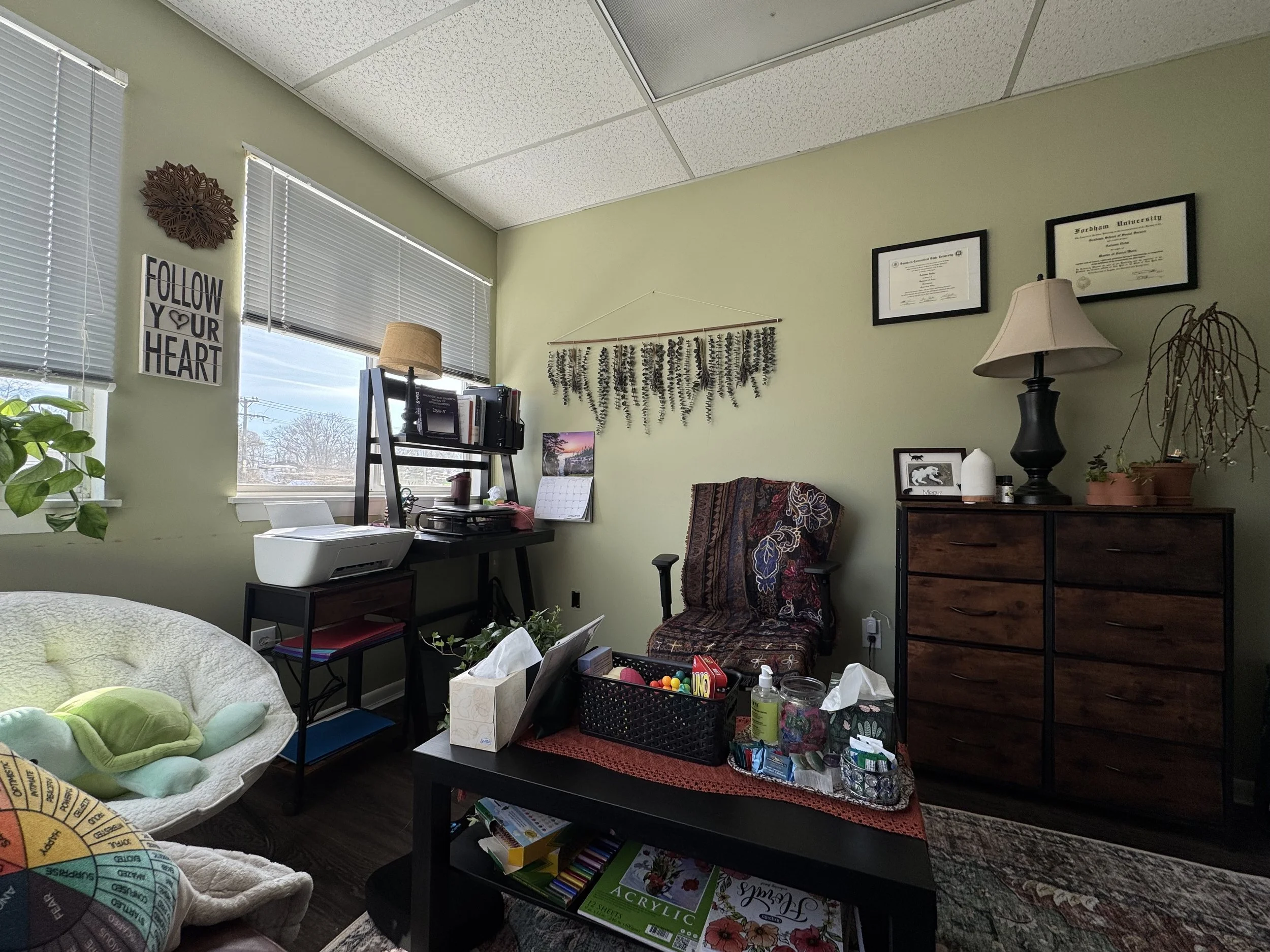 Cozy home office with a window, bookshelf, potted plants, armchair, dresser, lamp, wall decorations, and various office supplies.