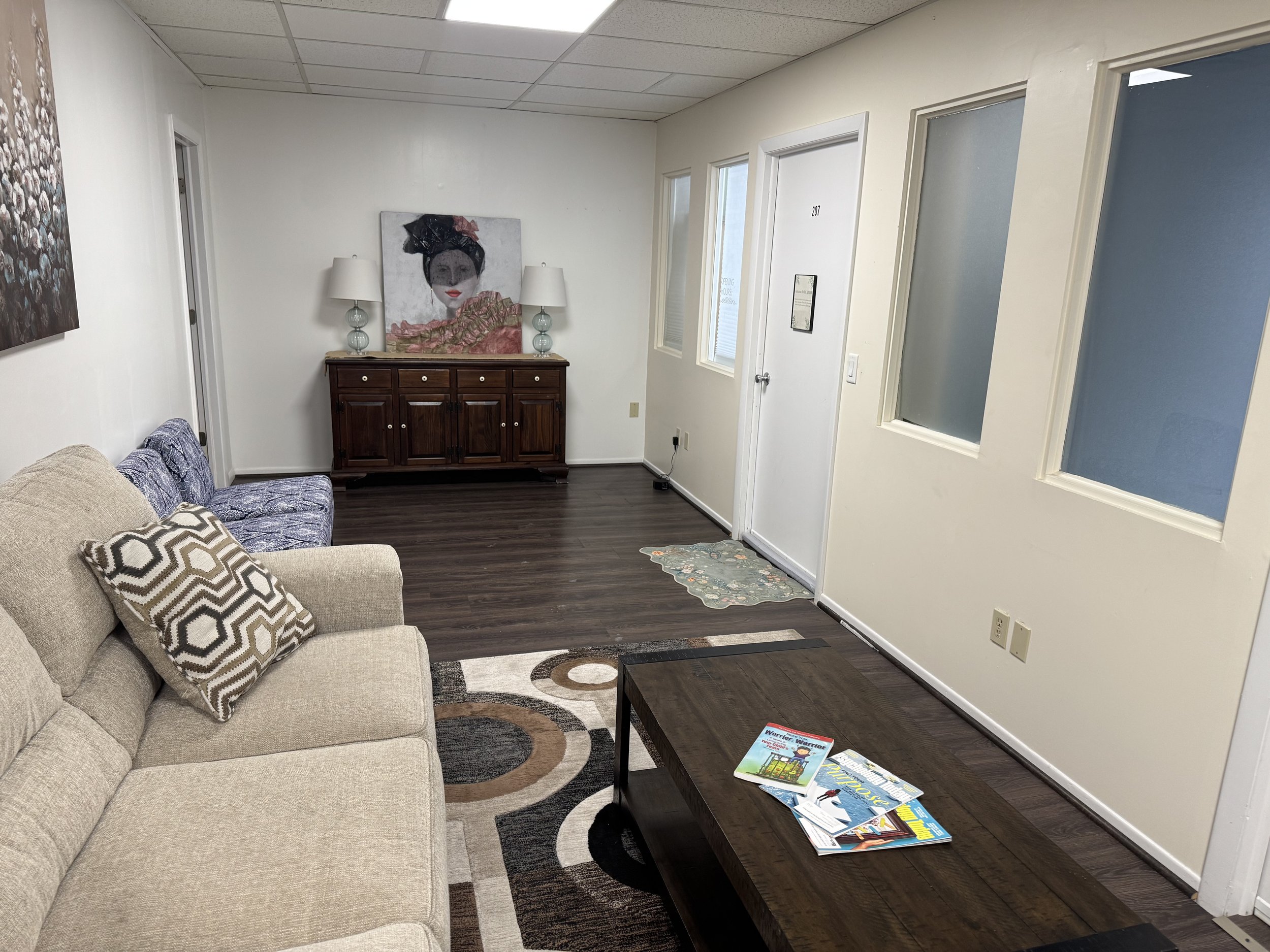 Waiting room with beige sofa, patterned pillow, blue armchair, wooden coffee table with magazines, dark wood floor, off-white walls, art on the wall, white door with a sign.