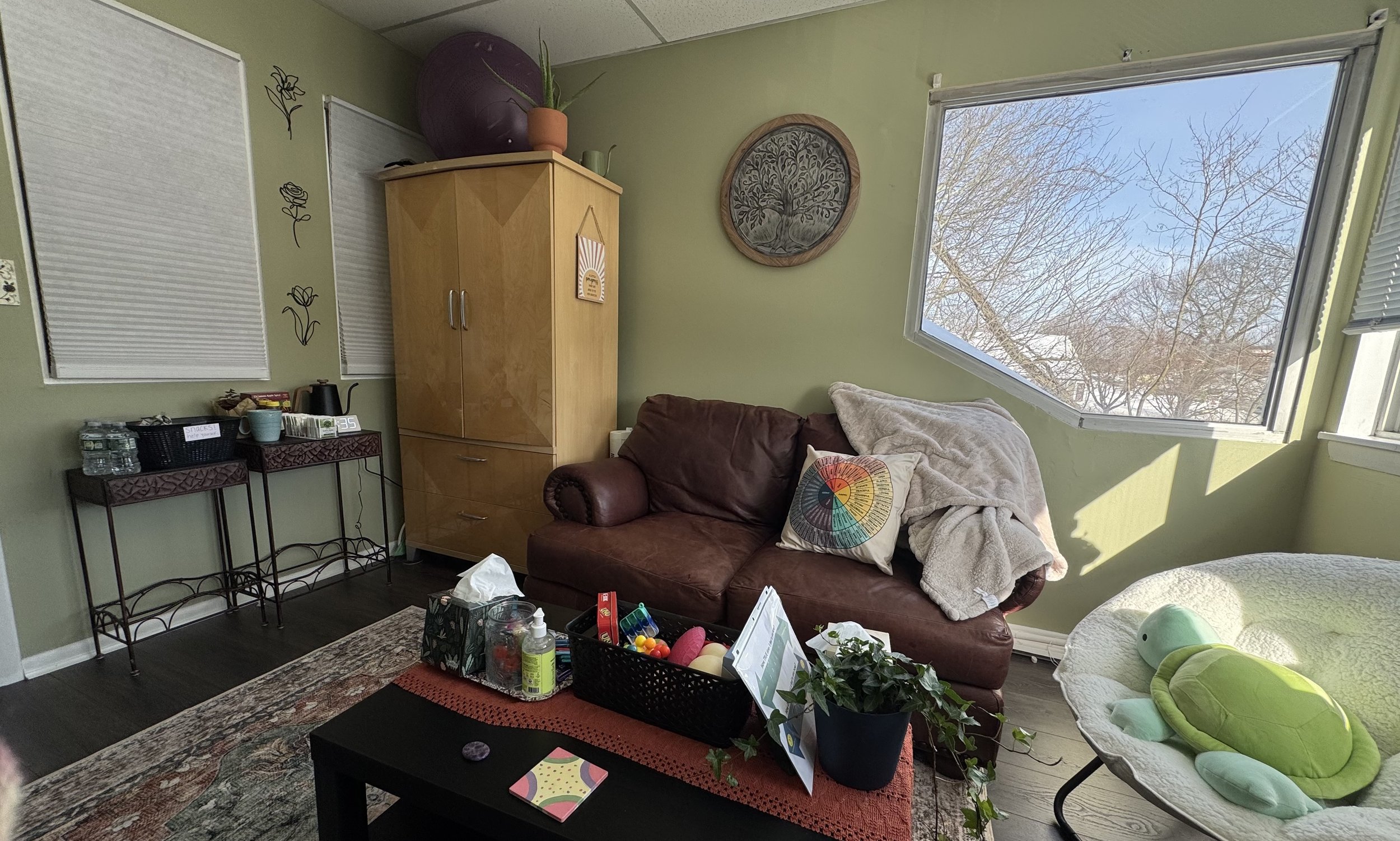 Living room with a brown leather sofa, a beige blanket, decorative pillow, and a round window showing snow-covered trees outside. A small table with toys and plants, a wooden cabinet, two small tables with water bottles and a kettle, and a white chair with a turtle pillow are also visible.