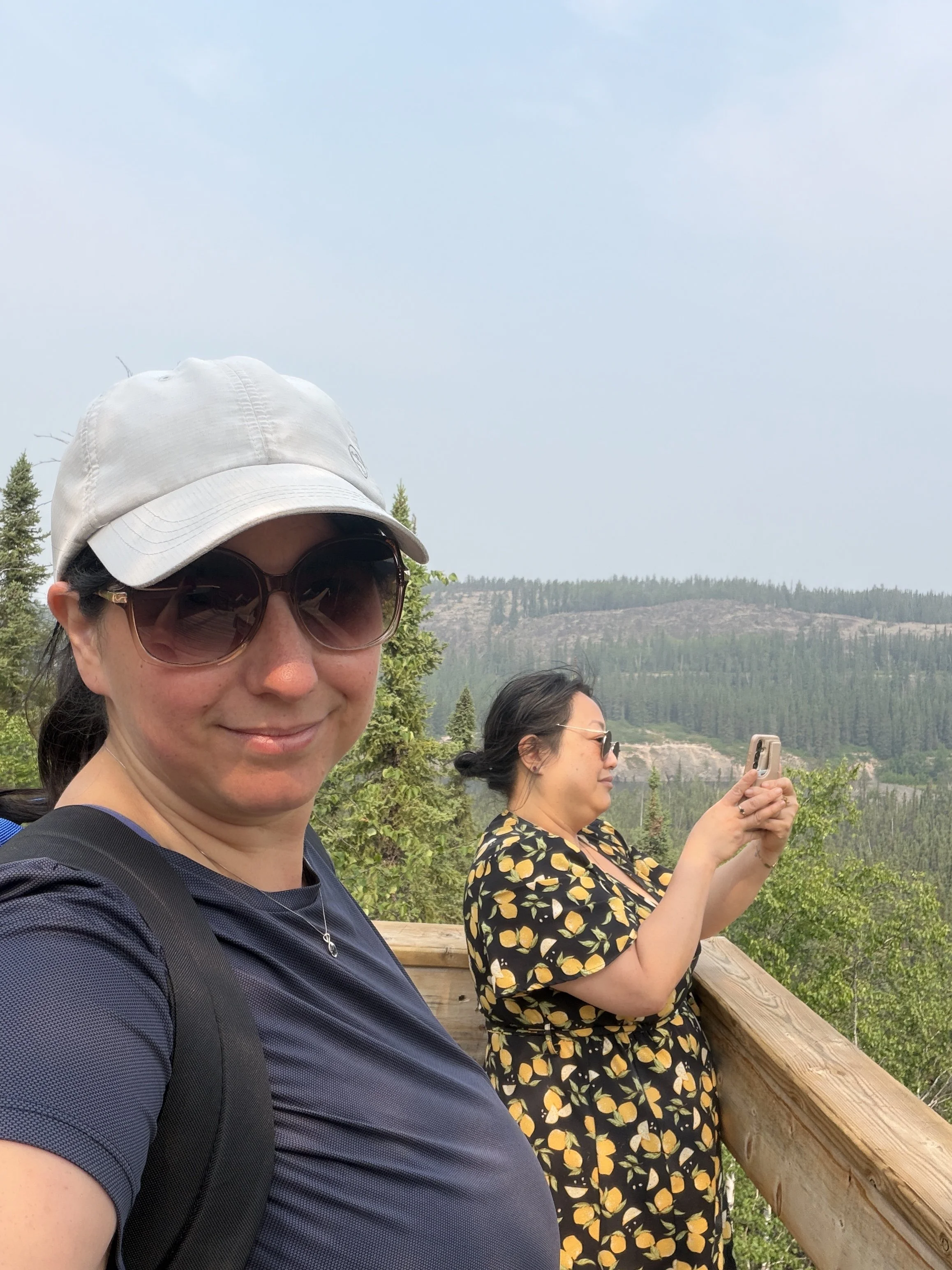 Two women at an outdoor scenic location with trees, hills, and a cloudy sky in the background. One woman is wearing a white hat, sunglasses, and a black shirt, smiling at the camera. The other woman is wearing glasses and a black dress with a lemon p
