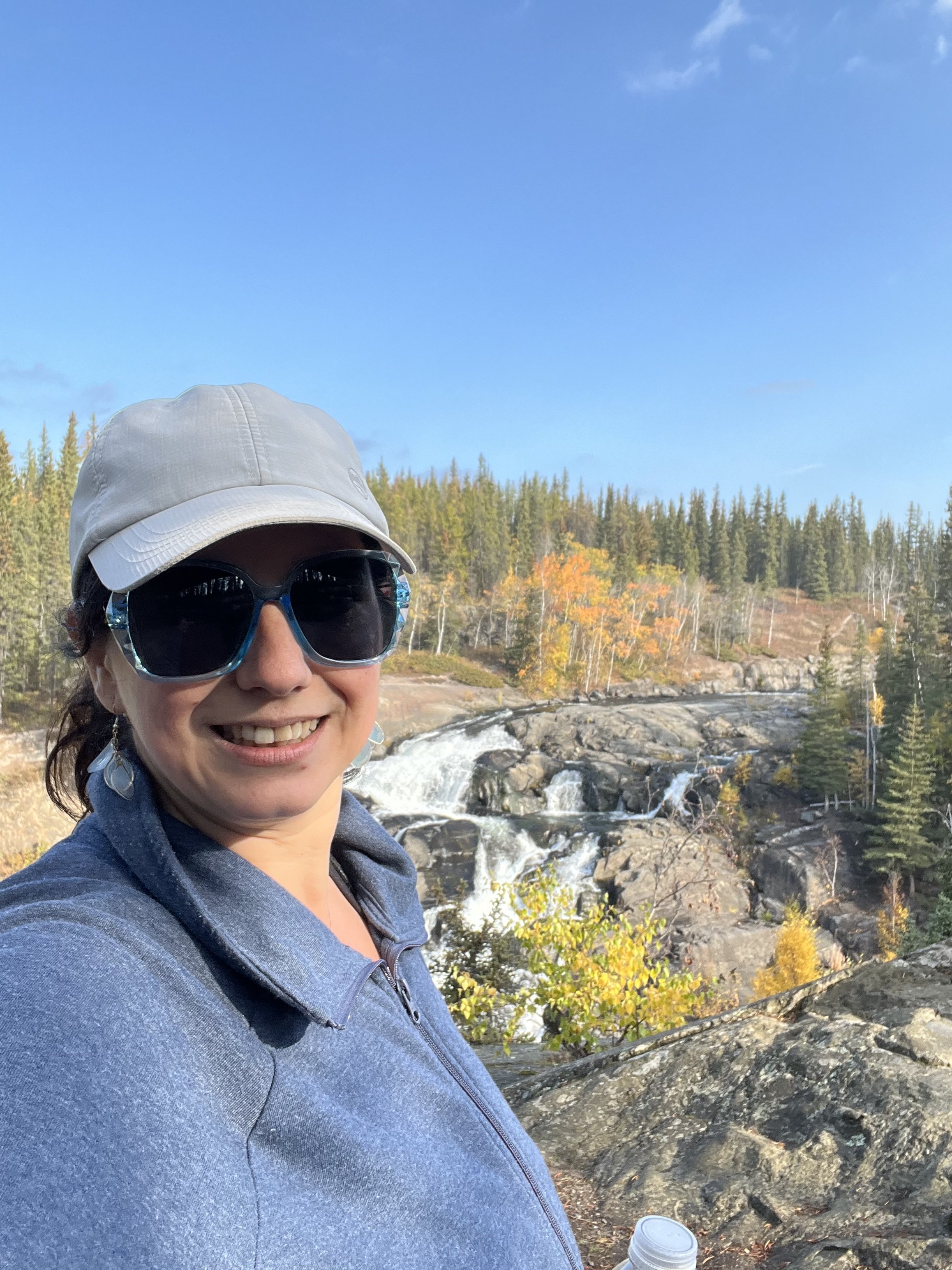 A smiling woman wearing sunglasses, a white baseball cap, and a gray hoodie takes a selfie outdoors near a river with rocks, trees with fall foliage, and a blue sky in the background.