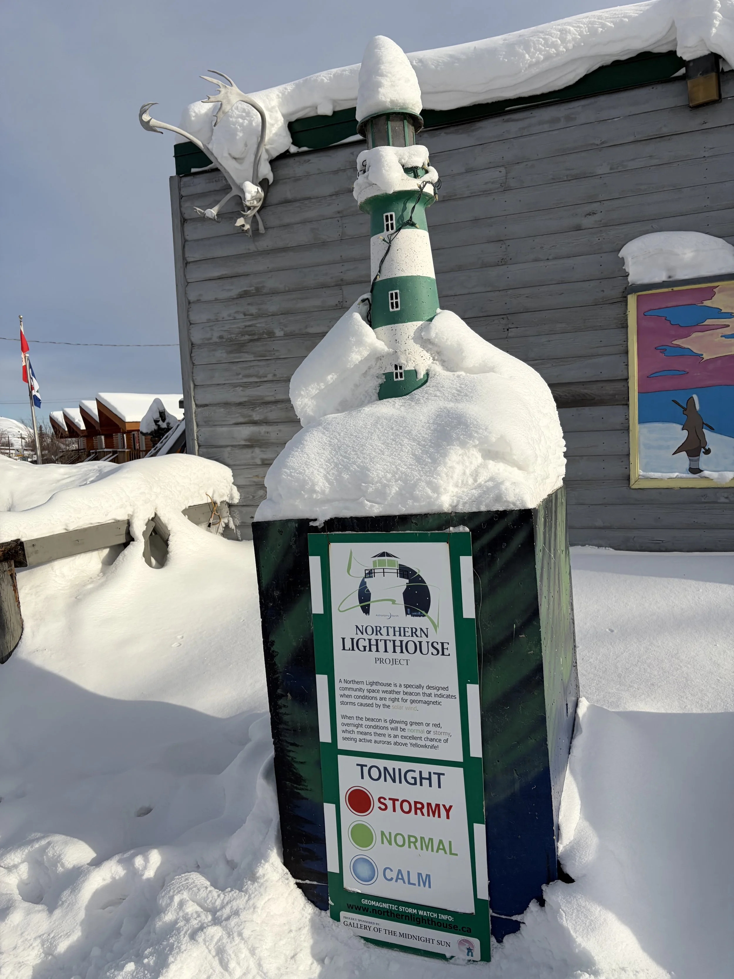 A snow-covered signboard reads 'Northern Lighthouse Project' and displays stormy weather conditions with color indicators for stormy, normal, and calm. It is placed outdoors in a snowy landscape with a wooden building in the background and snow on th