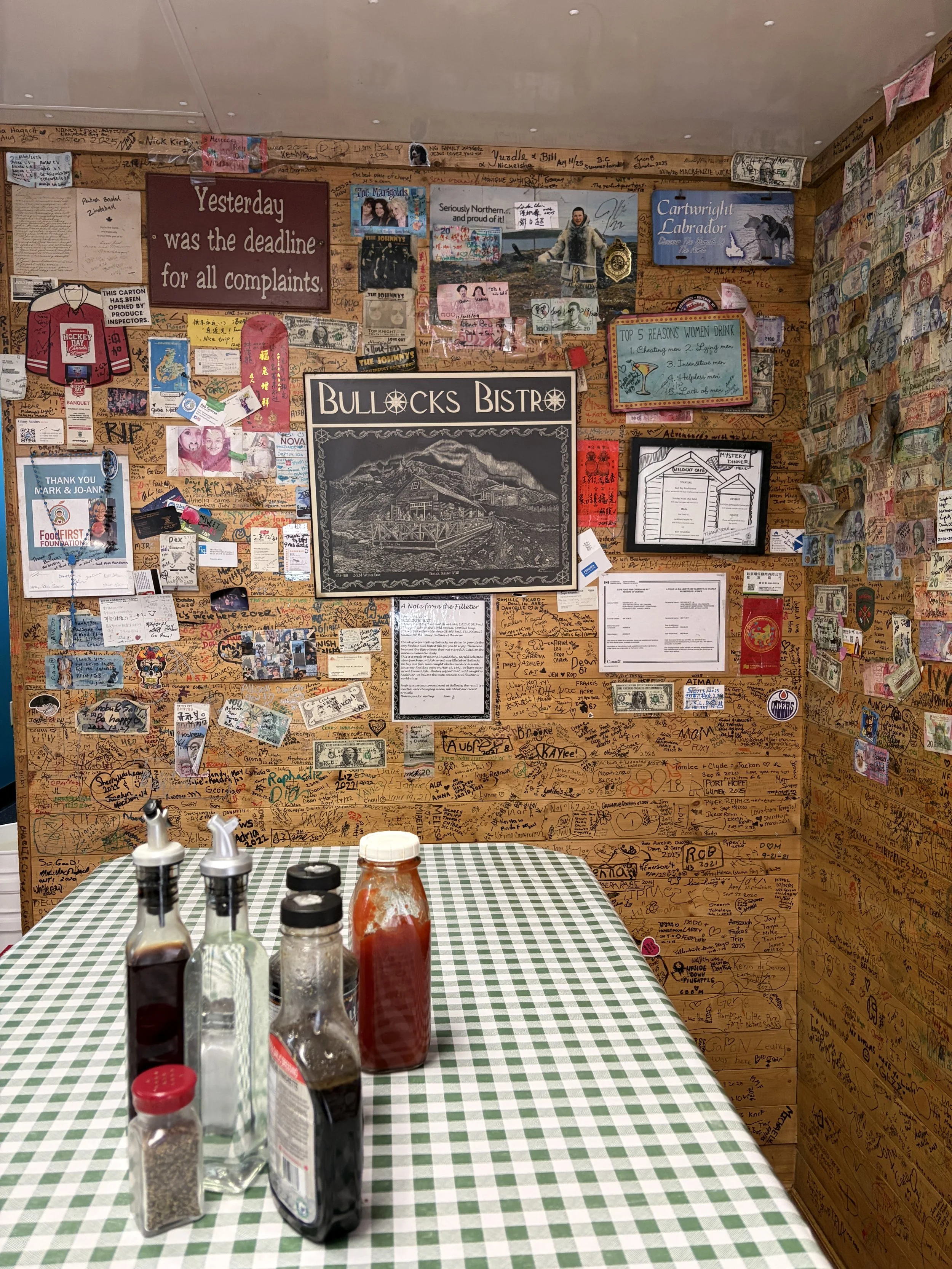 A table with a green and white checkered tablecloth. On the table are four bottles of condiments, including ketchup and soy sauce. The background wall is covered with handwritten messages, signatures, and various posters, including a large poster tha