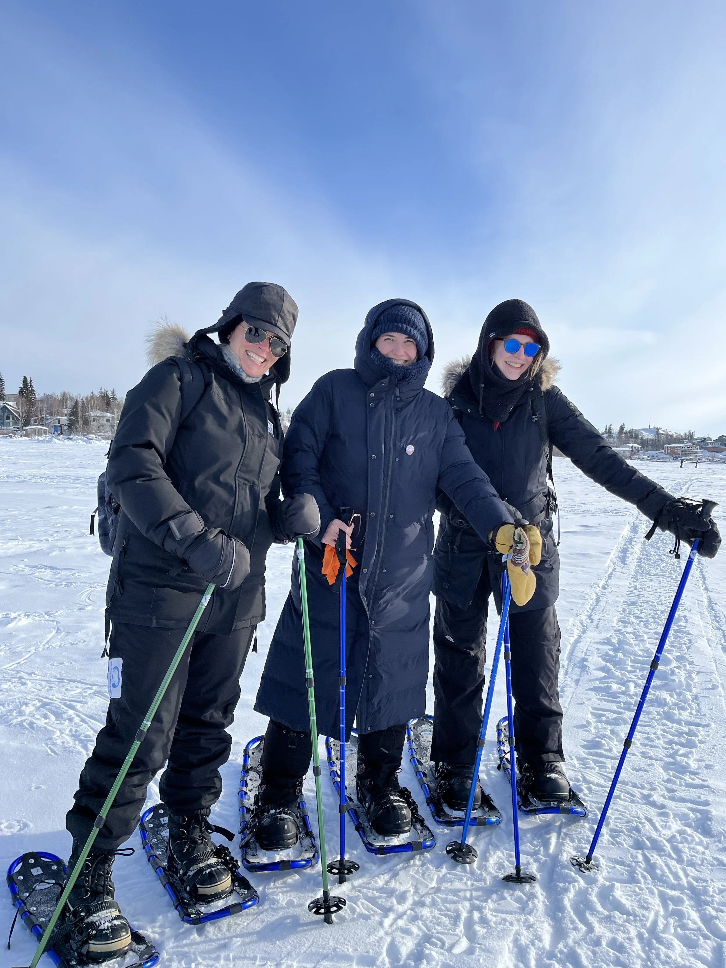 Three people in winter clothing standing on snow with snowshoes, holding trekking poles, and smiling at the camera against a clear blue sky with snowy houses in the background.