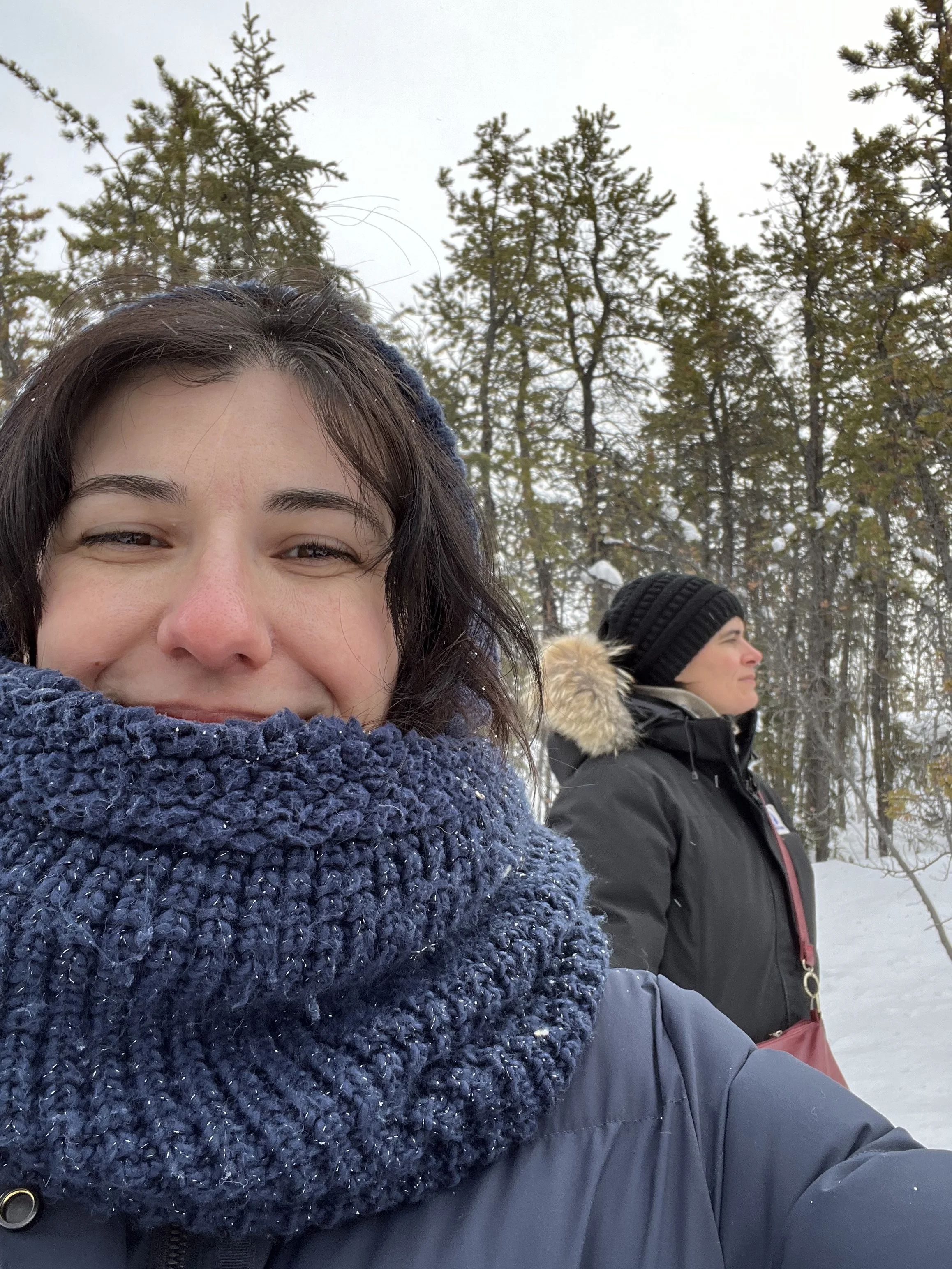 Close-up of a woman with dark hair and a blue scarf smiling outdoors in winter, with trees and snow in the background. Another person with a black beanie and winter coat is visible behind her.