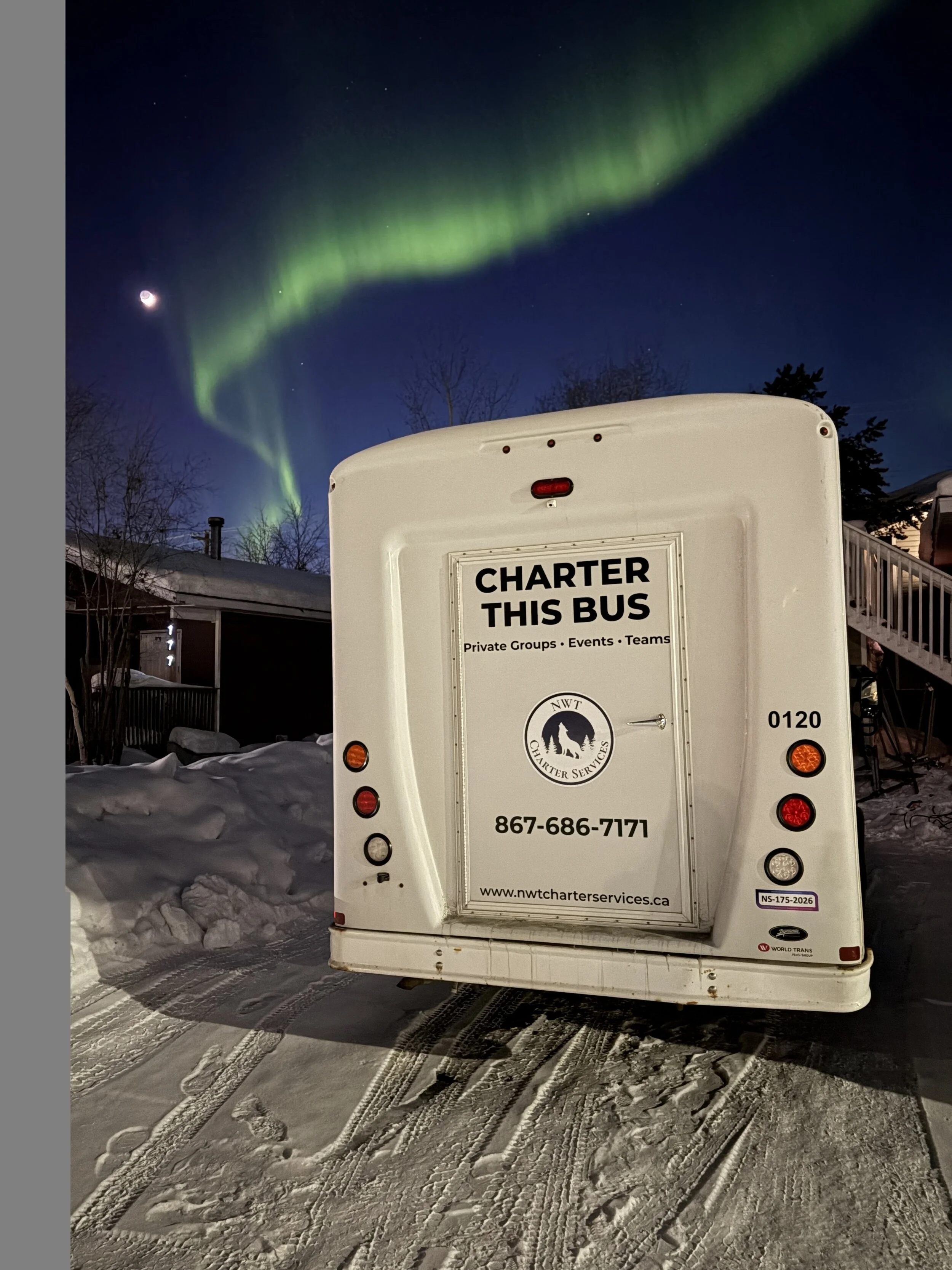 A white bus with the sign 'Charter This Bus' and contact information on the back, parked on a snowy street at night. The northern lights are visible in the sky above.