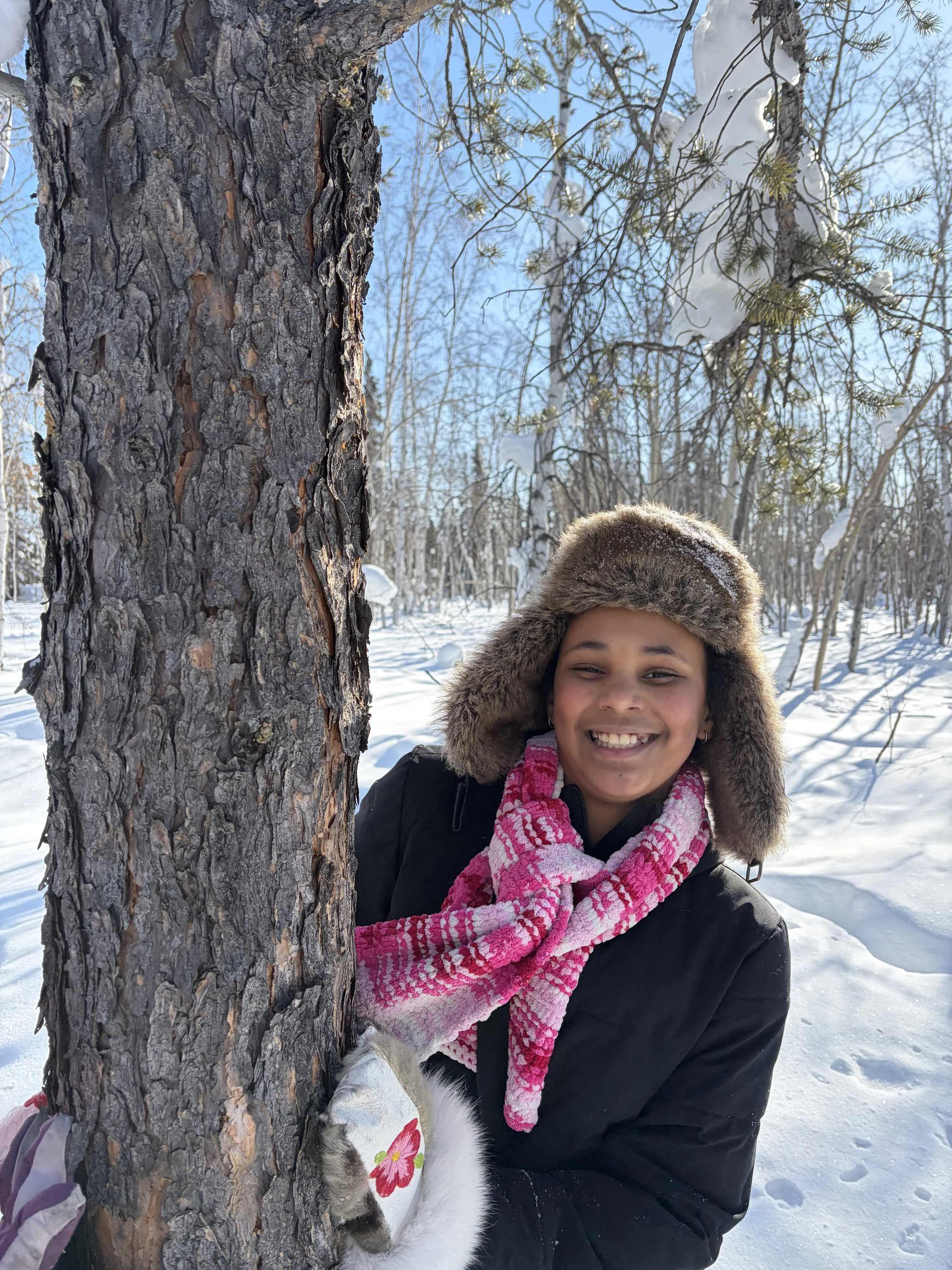 A young girl with brown skin, smiling, wearing a fur hat and pink scarf, standing outdoors in the snow, partially hiding behind a large tree trunk, with snow-covered trees and a clear blue sky in the background.