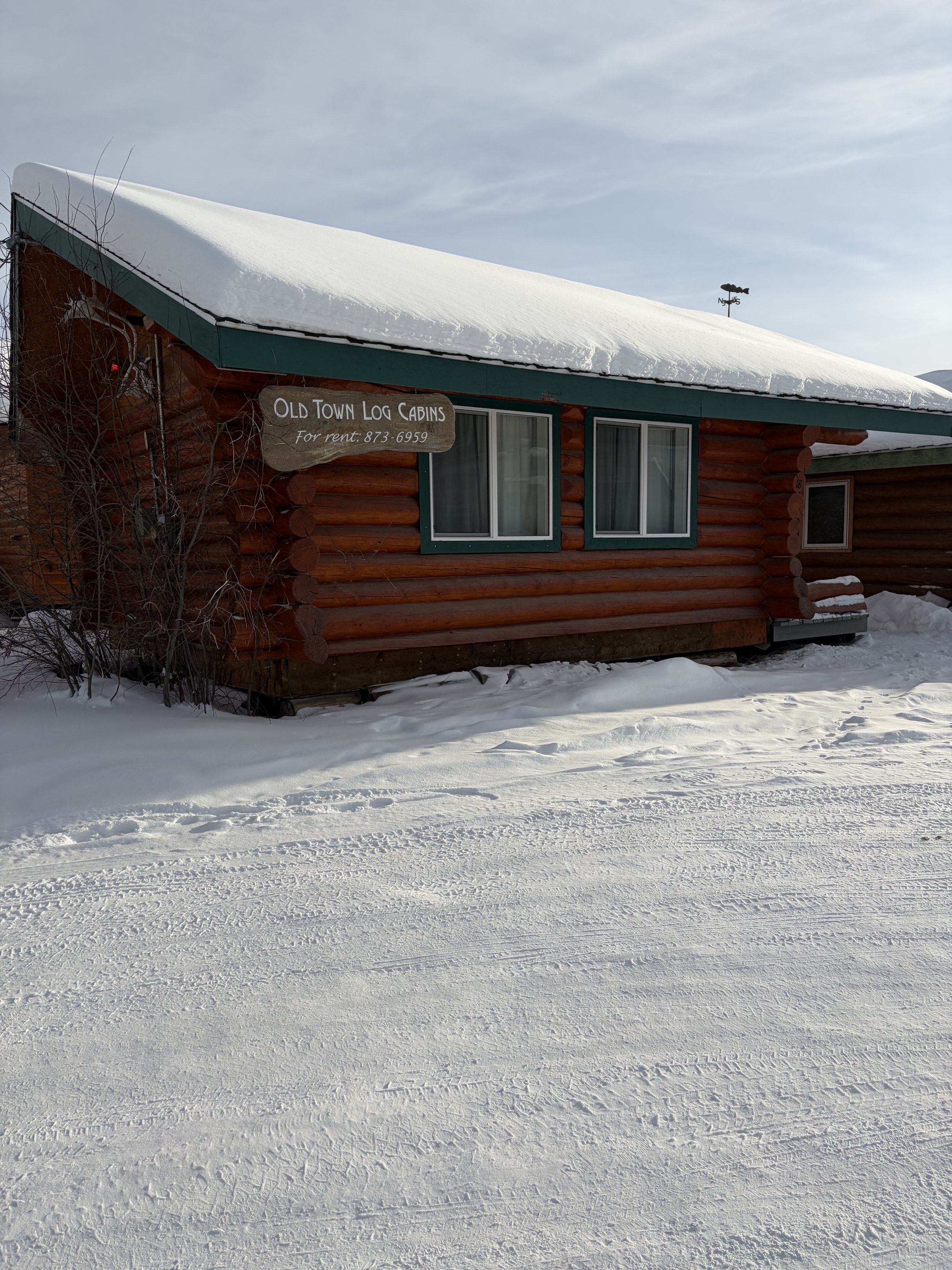 A log cabin with a snowy roof and a sign that reads 'Old Town Log Cabins for rent: 873-6959'. Snow covers the ground and the sky is partly cloudy.