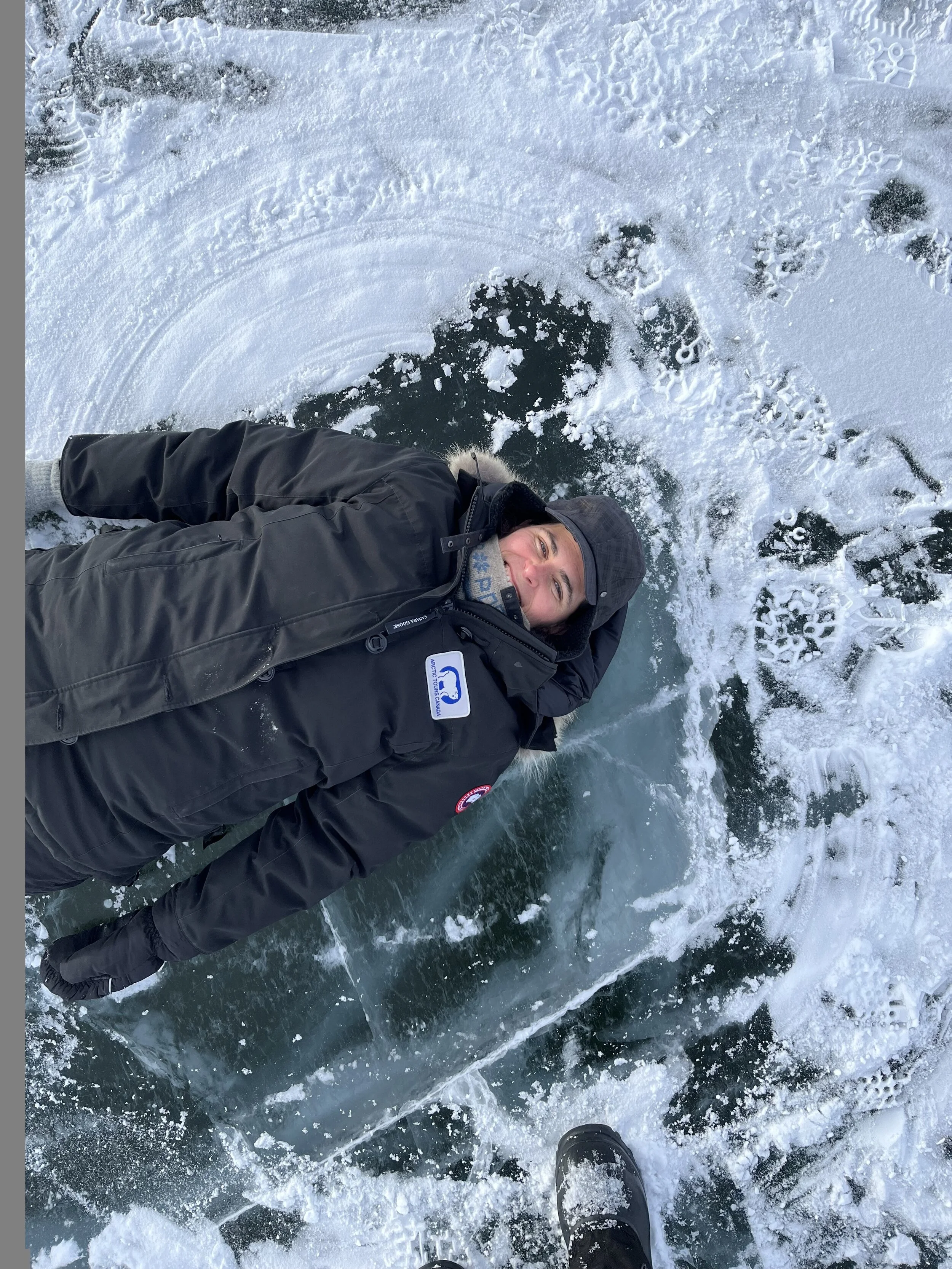 woman lying on an ice road with snow and ice around, wearing a black winter jacket and hat, with a happy expression.
