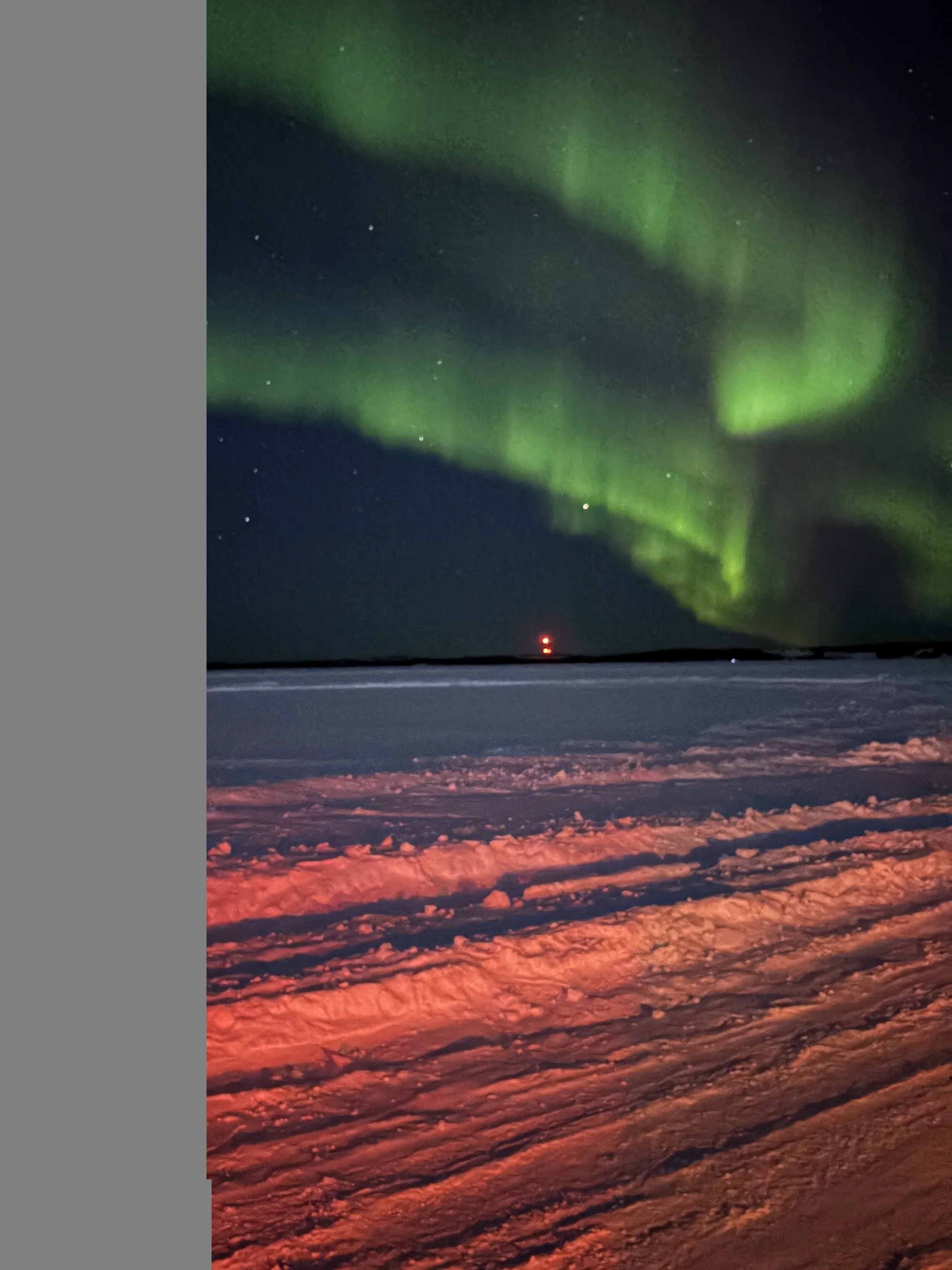 Night sky illuminated by the northern lights with green hues, above a snow-covered landscape with a road and a red warning light.
