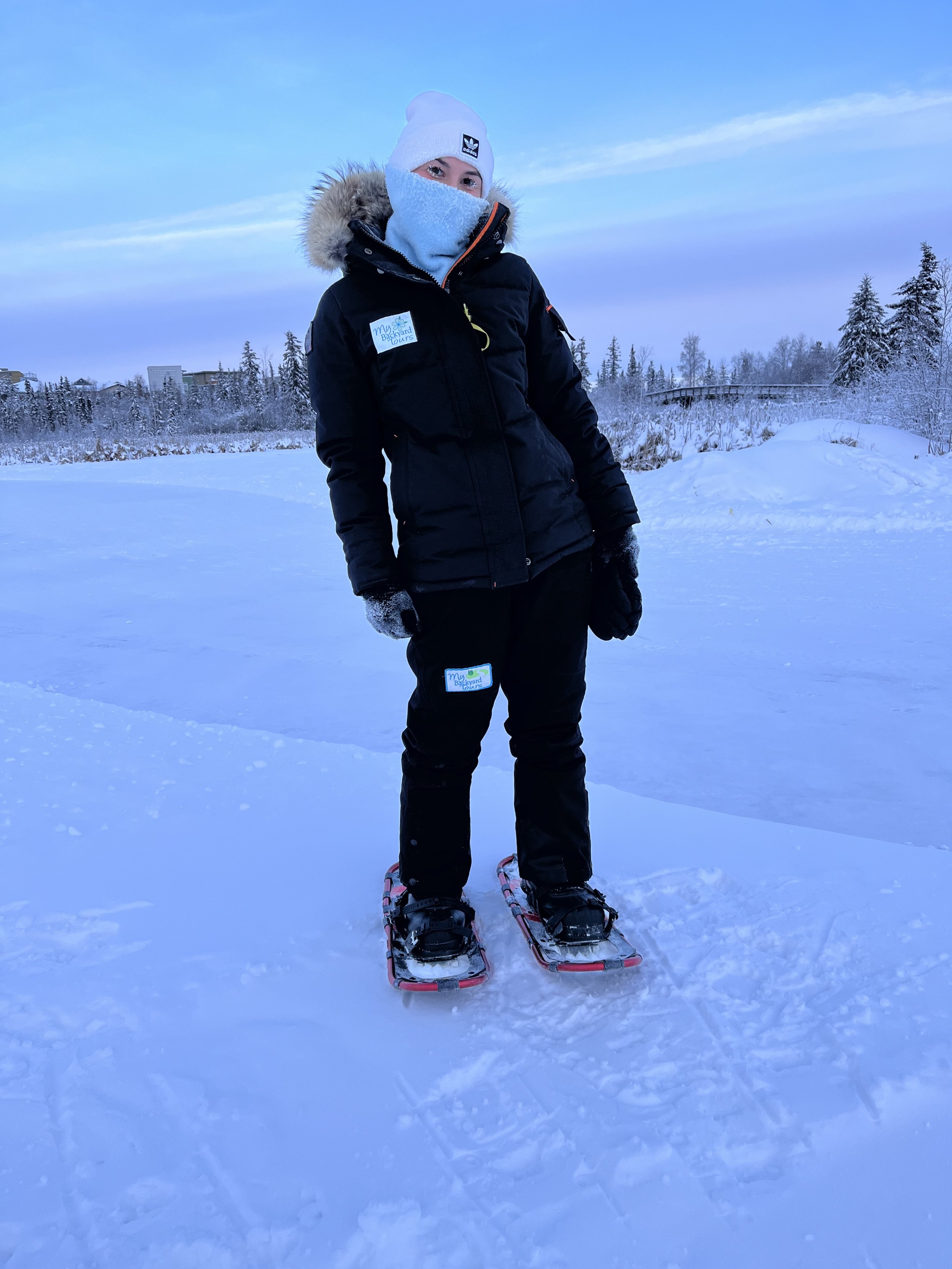 Person wearing a winter jacket, hat, and face covering, standing on snow-covered ground with snowshoes, in a snowy outdoor landscape with trees and buildings in the background.