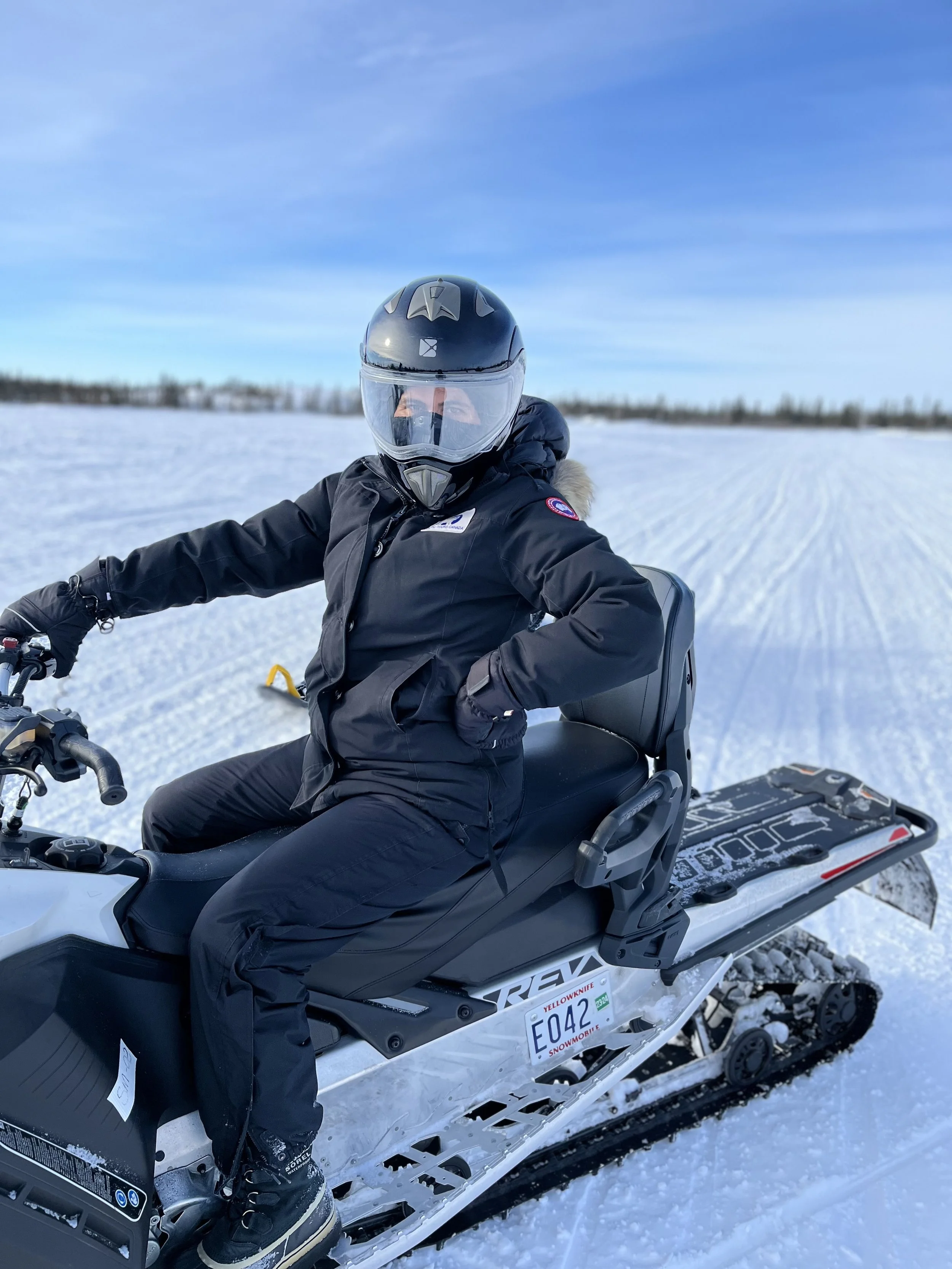 woman in black winter parka and helmet riding a snowmobile on snowy terrain under blue sky.