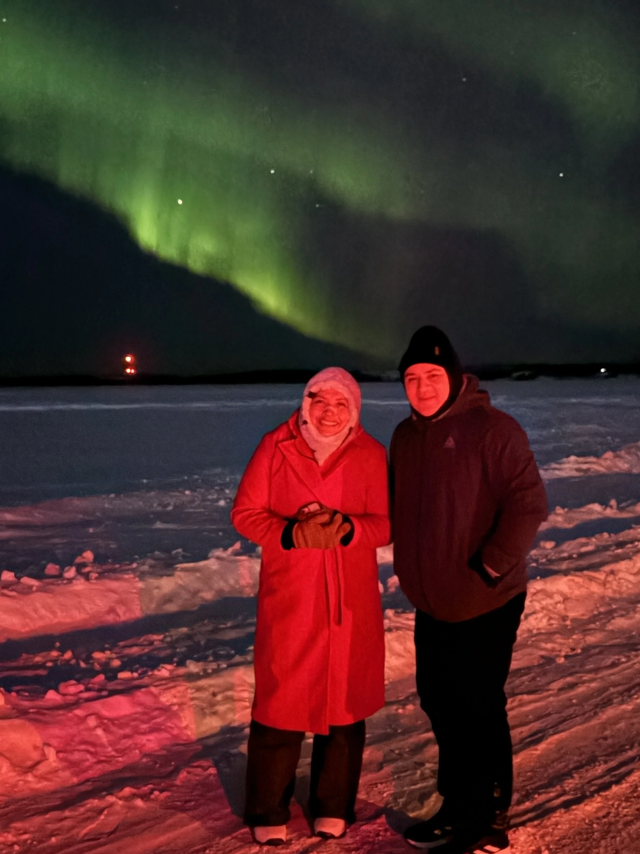 Two people standing on snow-covered ground at night, illuminated by red and green lights, with the Northern Lights visible in the night sky above.