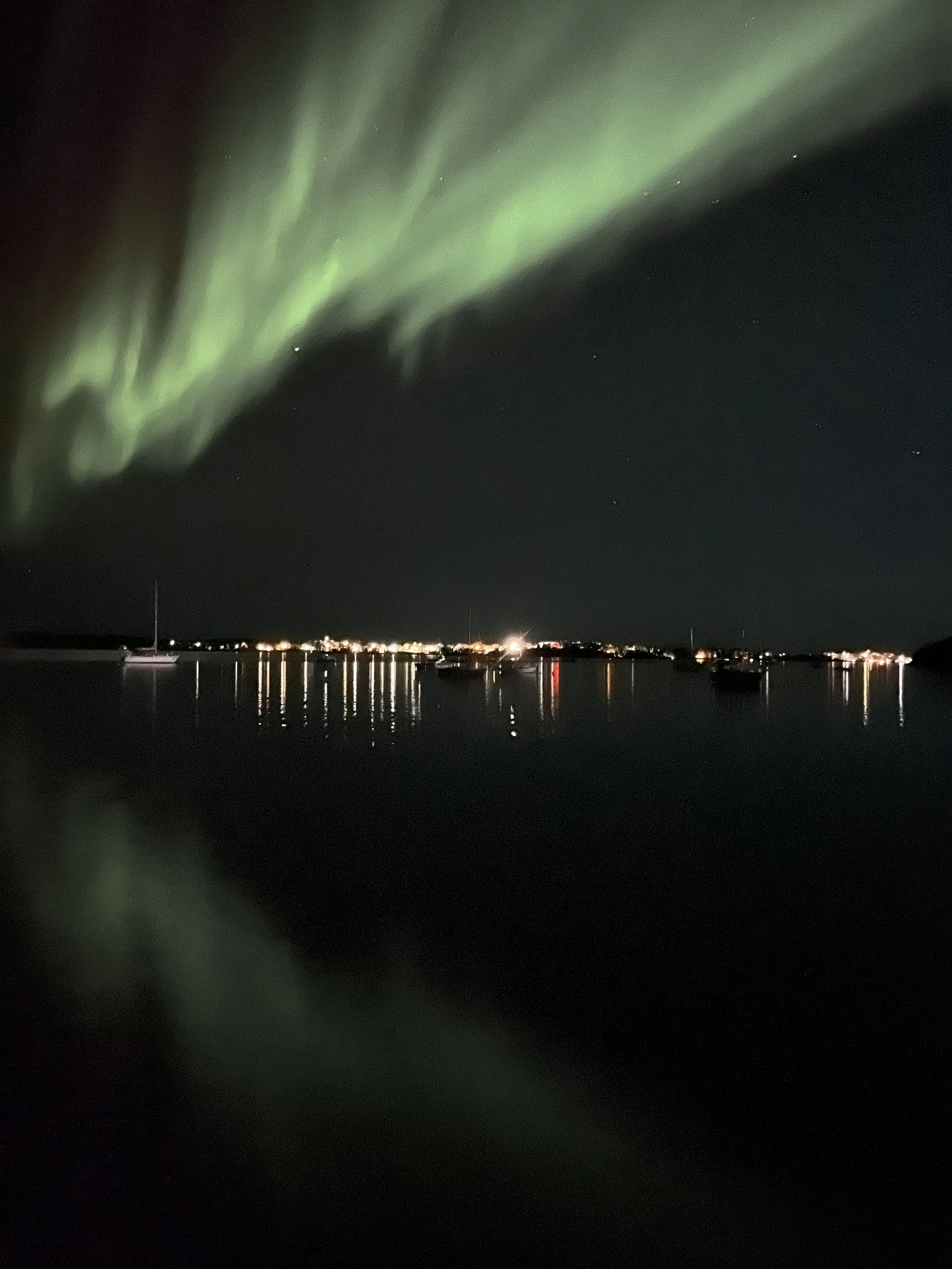 Northern lights (aurora borealis) over a dark body of water with boats and a distant shoreline with lights at night.