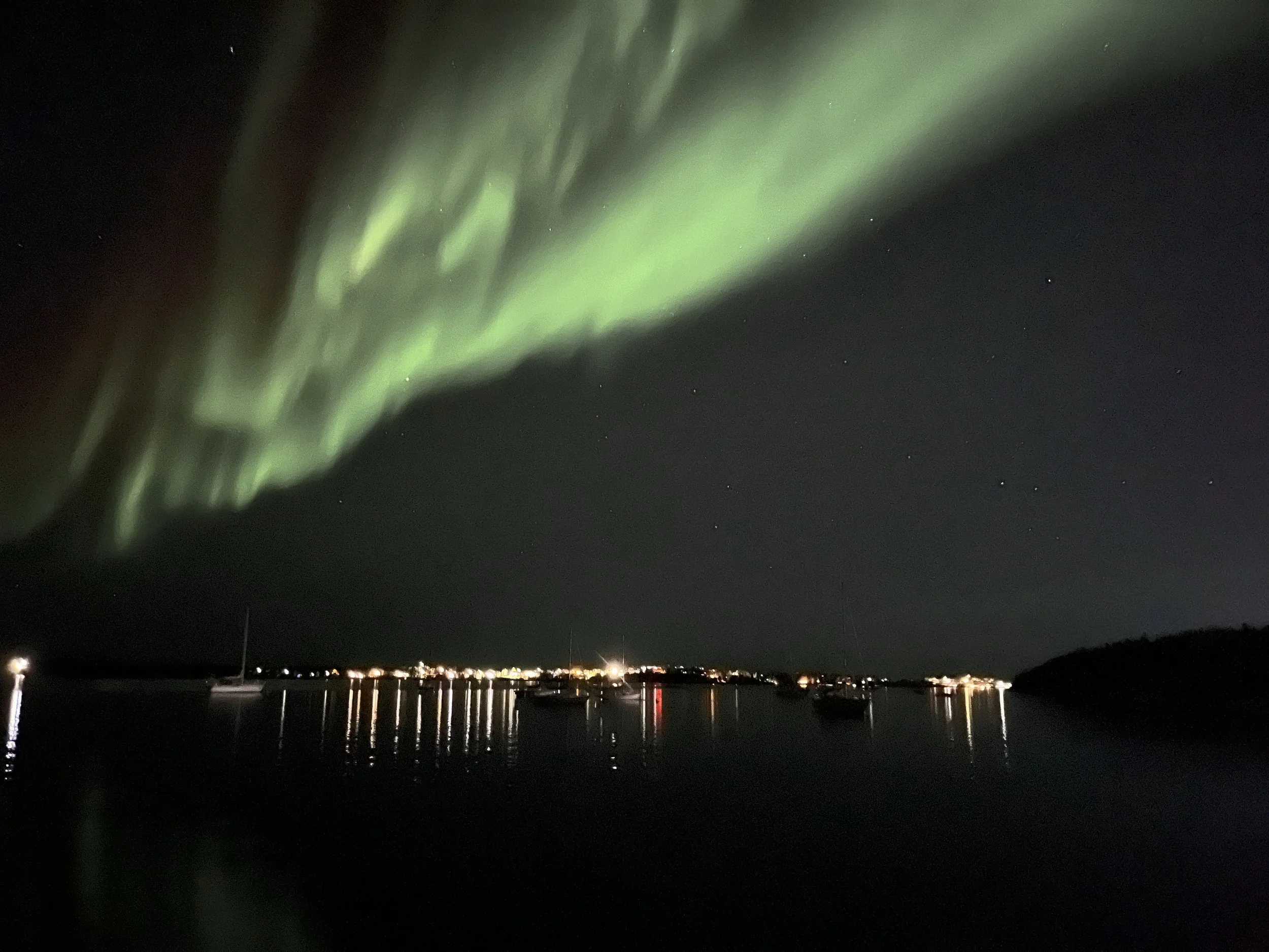 Nighttime scene of the northern lights (aurora borealis) with green light streaks across the sky, stars visible, and a body of water with boats and reflections below.