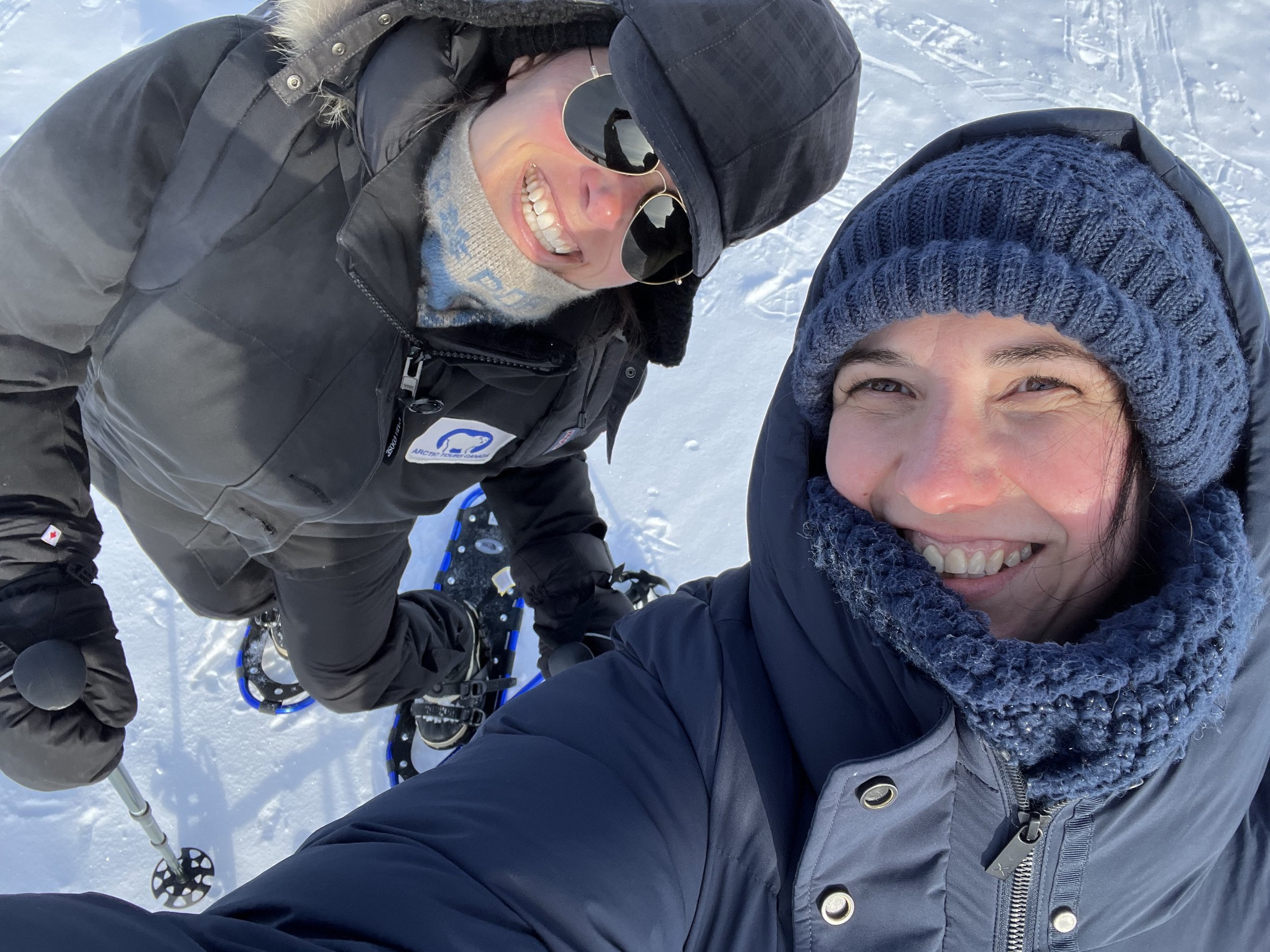 Two women taking a selfie on a snowy landscape, dressed warmly in winter clothing, including hats, scarves, and jackets, smiling and enjoying the outdoor winter activity.