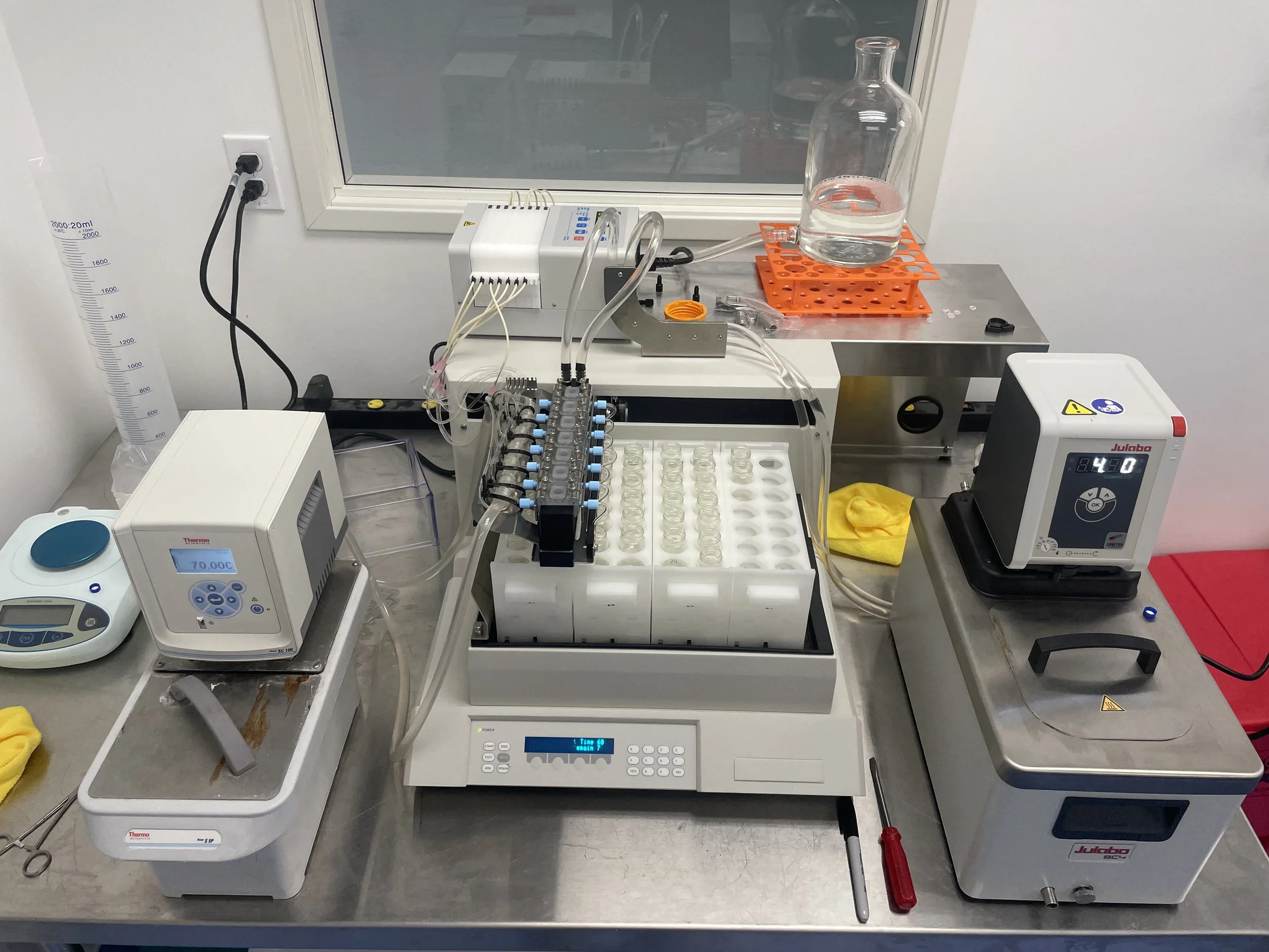 Laboratory setup with various scientific equipment including a glass bottle with liquid, test tubes, pipettes, and heating blocks on a stainless steel workbench. Franz cells. Permeation equipment. Diffusion. Fractionate. Permegear. Skin testing