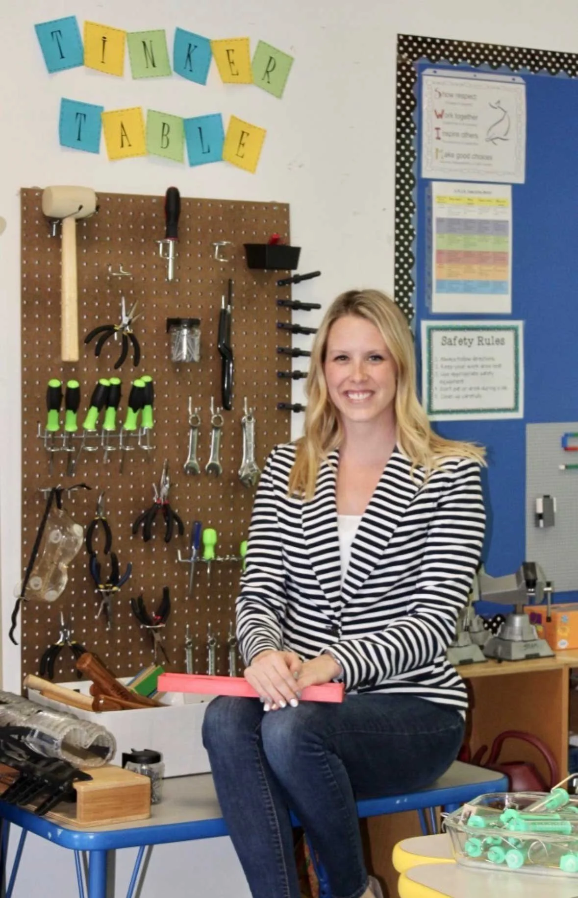 A woman sitting on a blue school chair in a classroom with a pegboard wall of tools behind her, including hammers, screwdrivers, pliers, and safety equipment, in front of a blue bulletin board with educational posters.