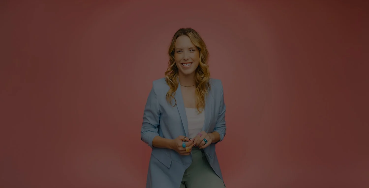 A woman smiling, wearing a light blue blazer, white top, and light-colored pants, standing in front of a pink background.