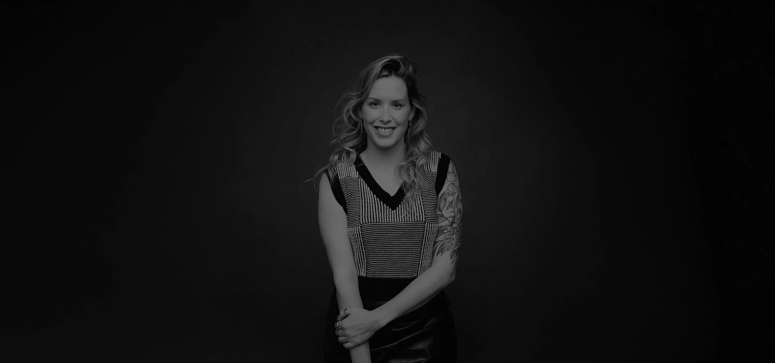 Black and white portrait of a woman smiling, with wavy hair, wearing a sleeveless striped top and a tattoo on her left arm, standing against a dark background.