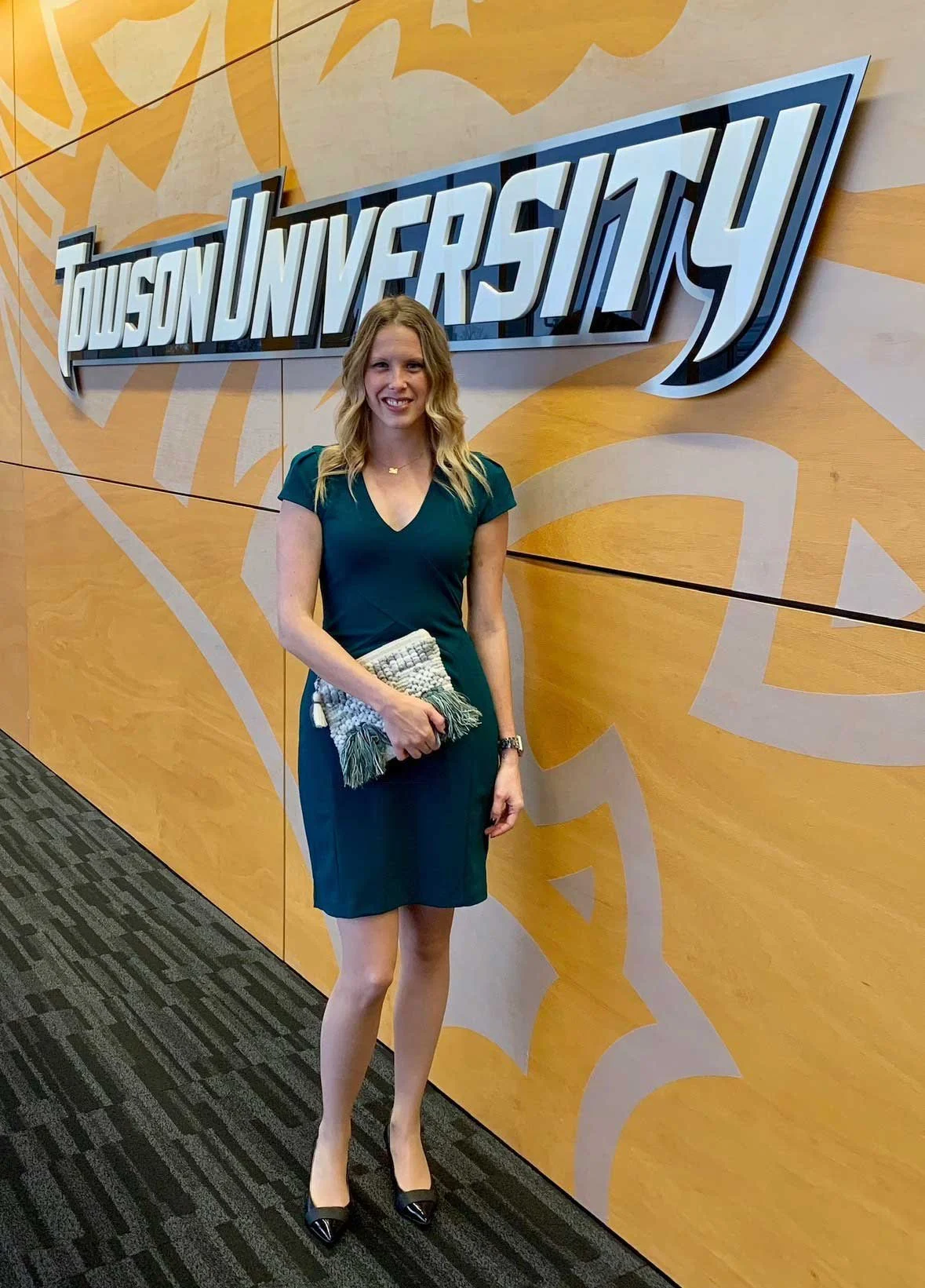 A woman standing in front of a wall with a "Dudson University" sign, wearing a dark green dress, holding a woven clutch, with long wavy blonde hair and black heels.
