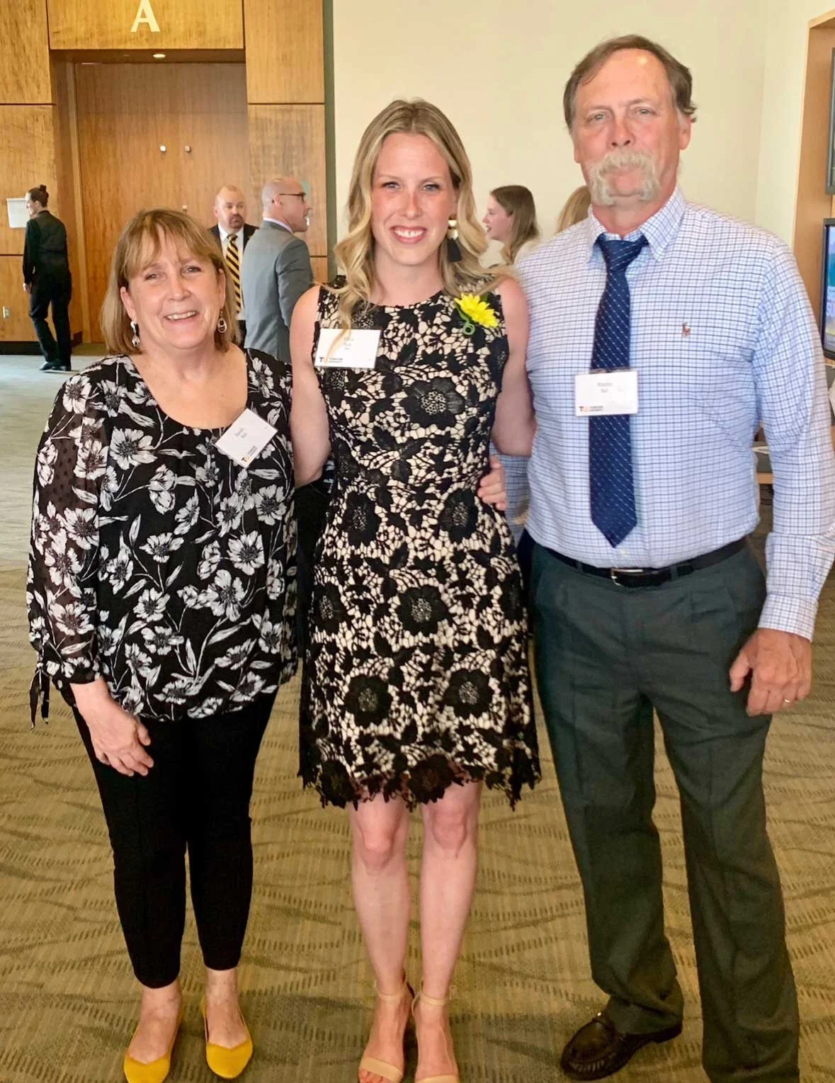 Three people standing together at a professional event, smiling for a photo. The woman in the middle is wearing a black and white floral dress with a yellow flower on her left shoulder. The woman on the left is dressed in a black and white floral top and black pants with yellow shoes. The man on the right is dressed in a light blue checked shirt, dark pants, and a blue tie.