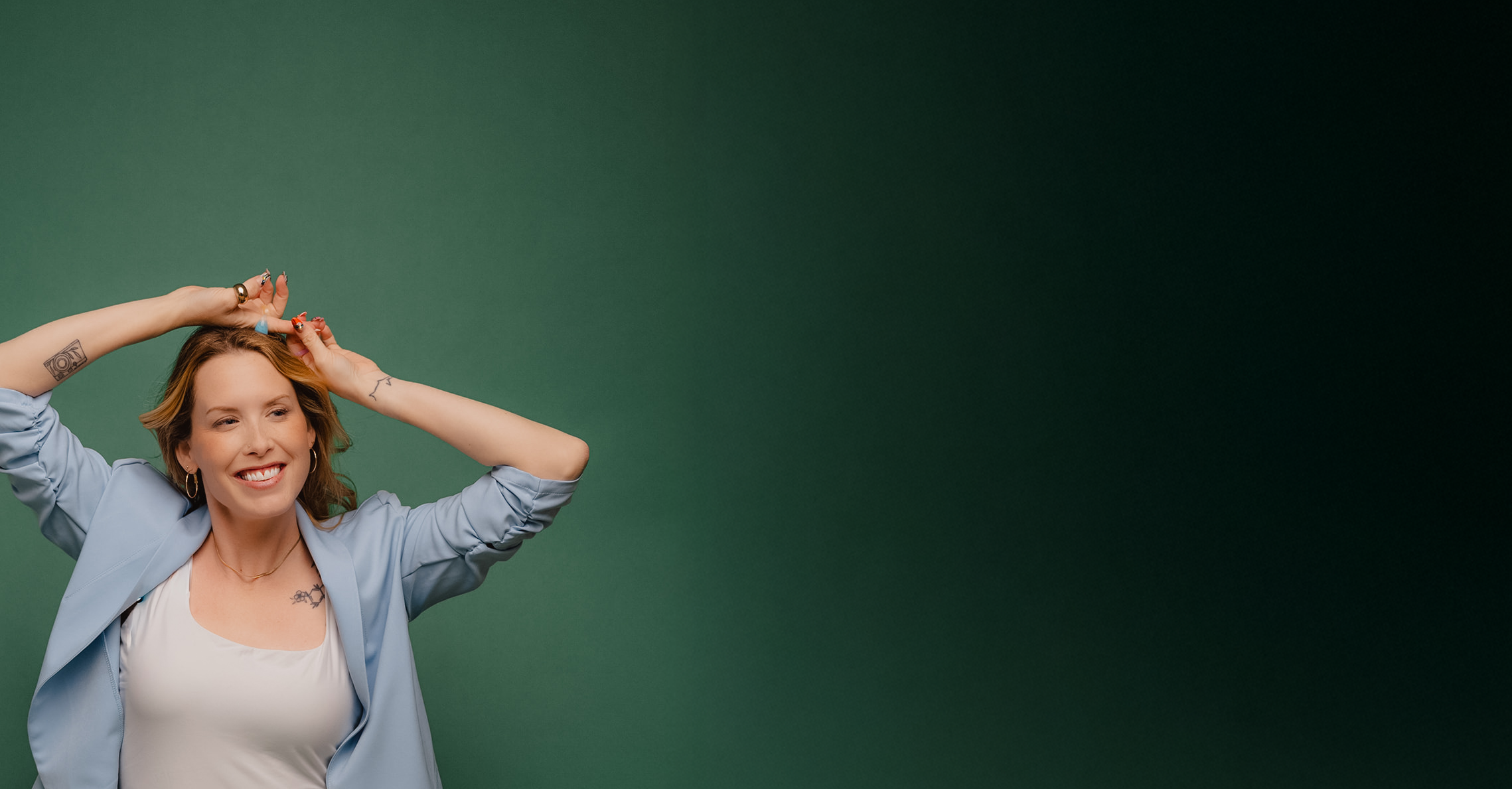 A woman with tattoos on her arms, wearing a light blue blazer and a white top, smiling and posing with her hands behind her head against a green background.