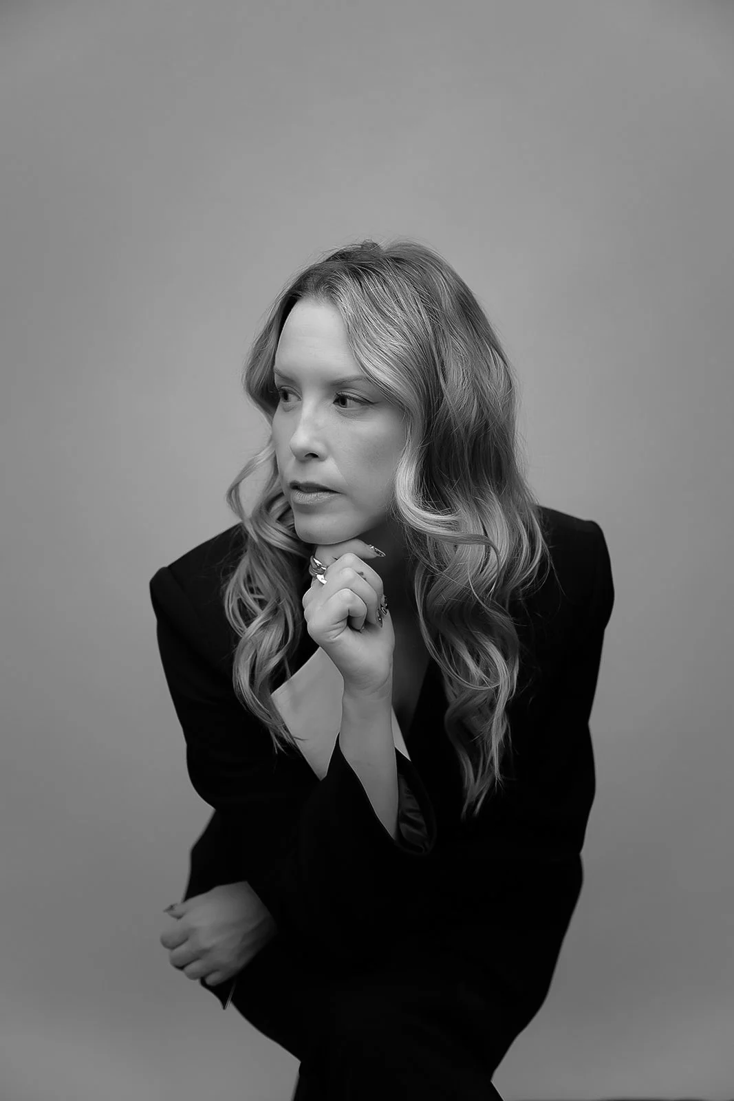 Black and white portrait of a woman with wavy blonde hair, wearing a dark jacket, looking to the side with her chin resting on her hand.