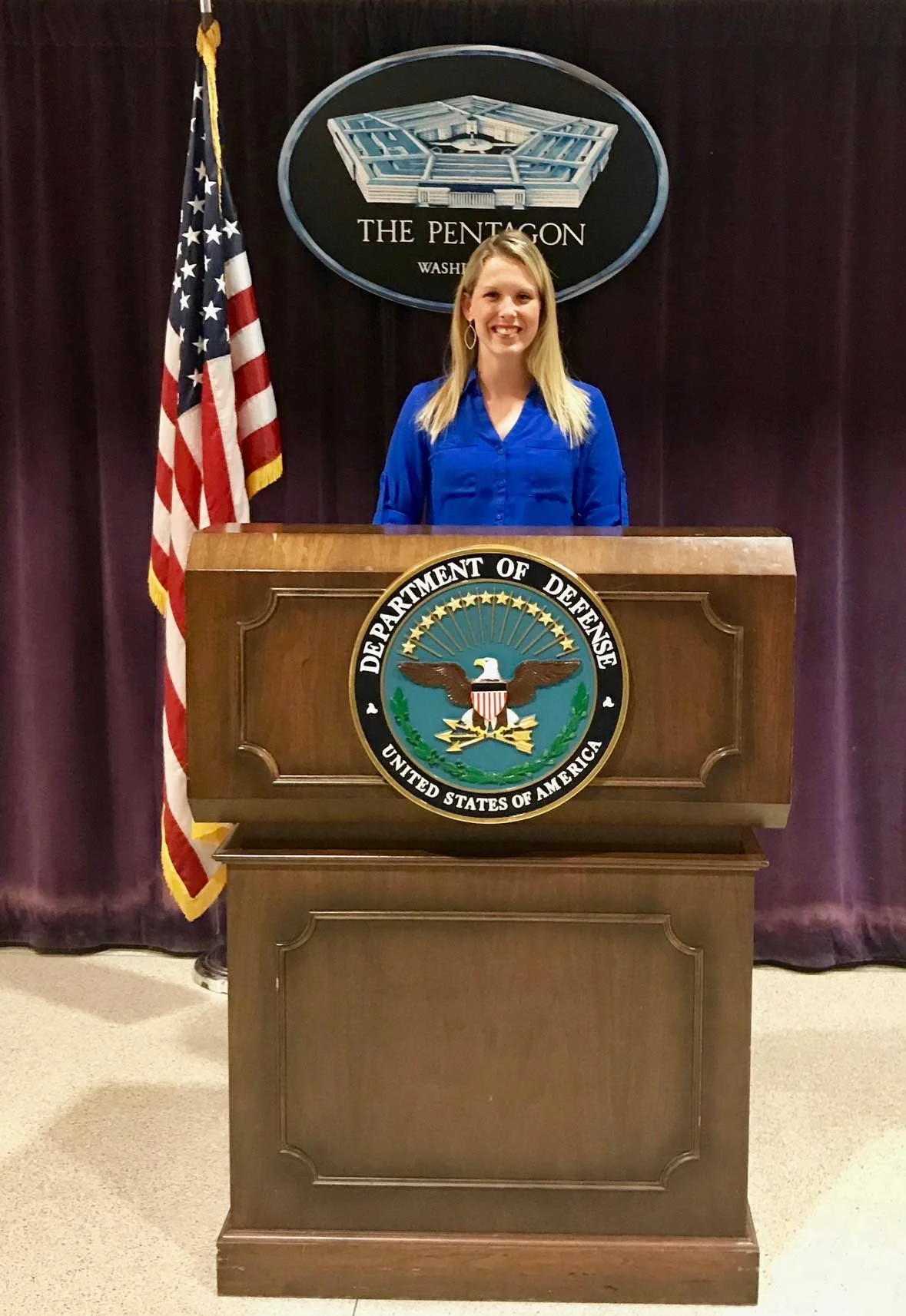 A woman standing behind a wooden podium with the Department of Defense emblem, in front of a dark curtain and an American flag, at the Pentagon in Washington, D.C.