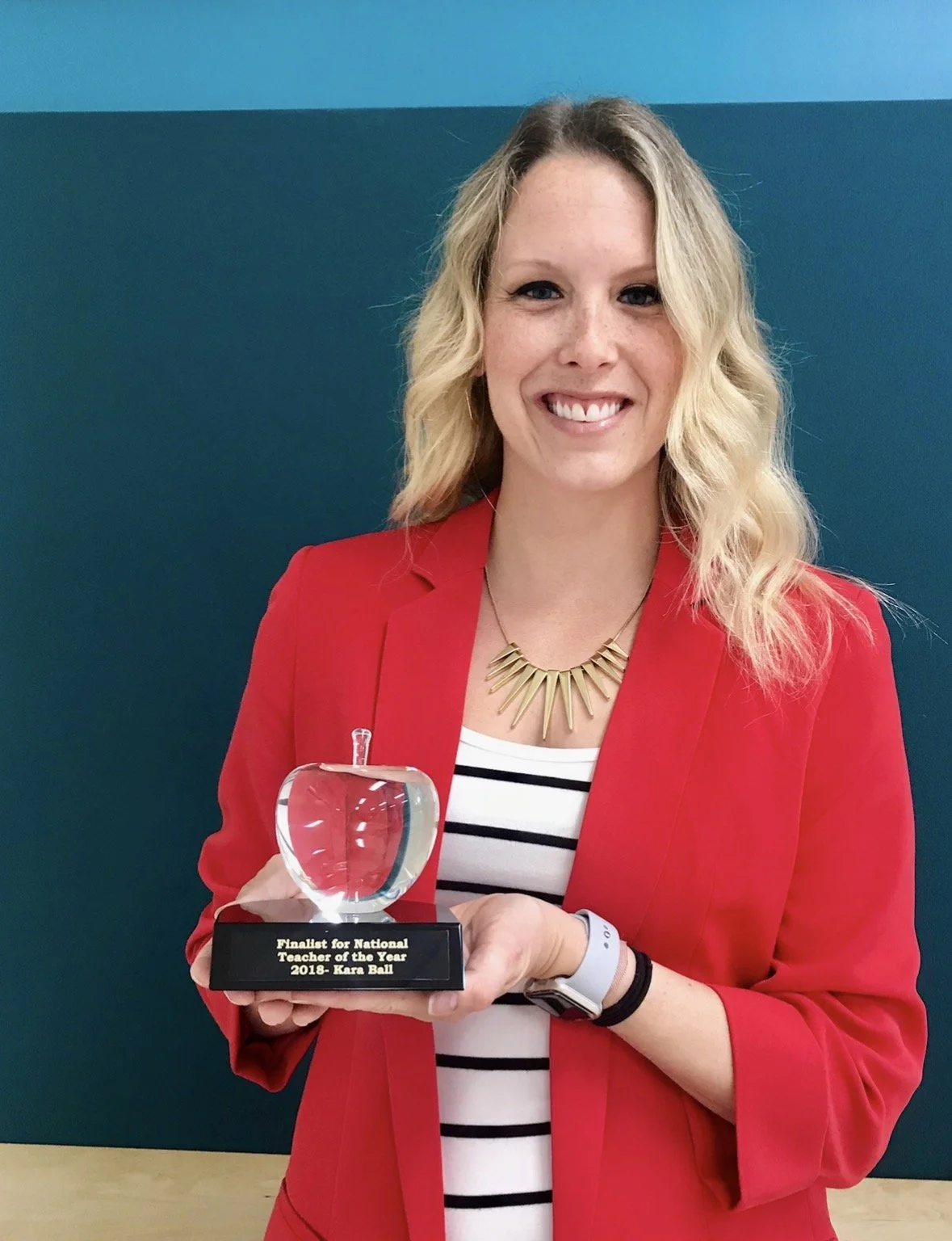 Woman in red blazer holding a glass apple trophy, smiling, with a blue-green wall in background.