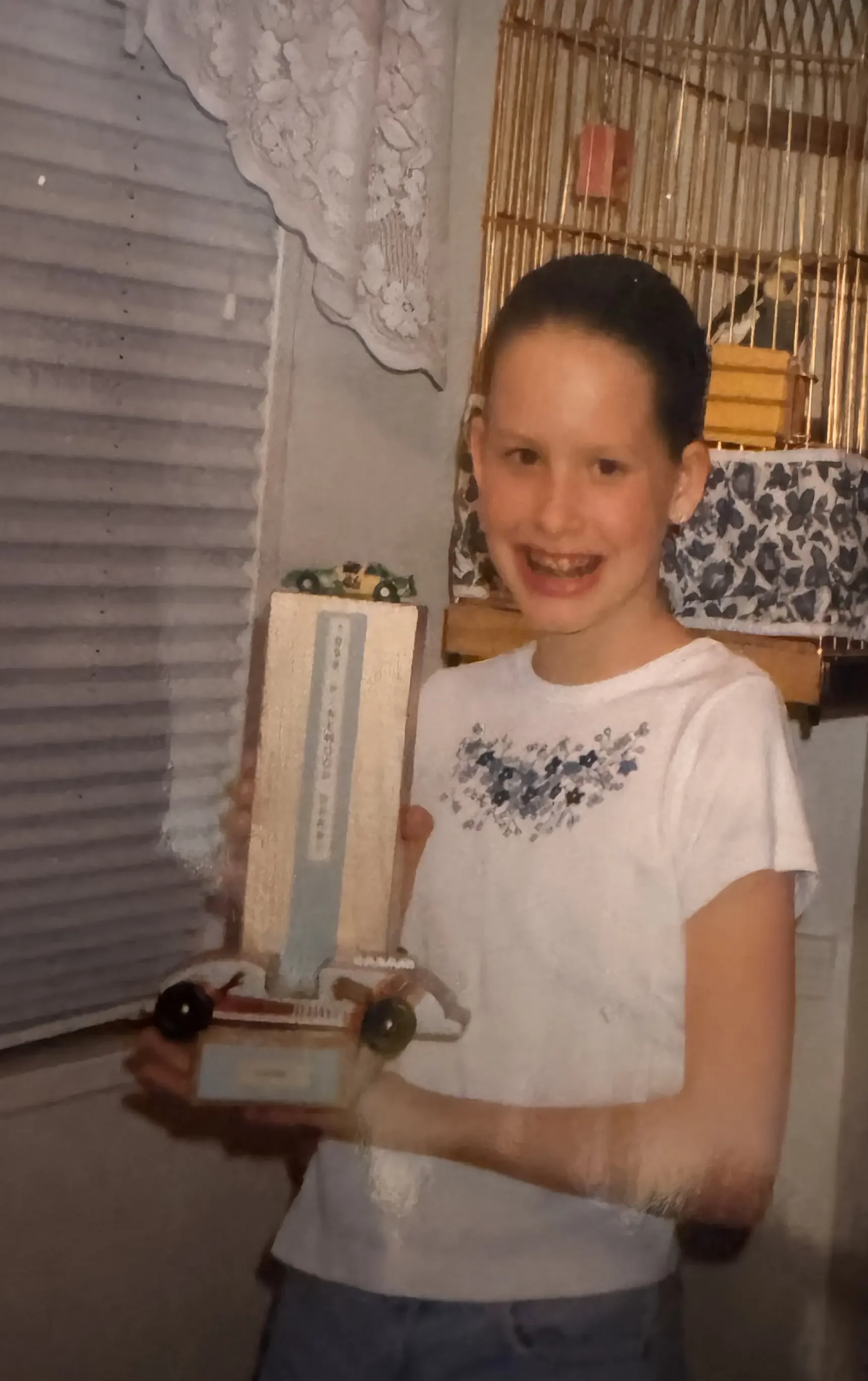 A young girl smiling and holding a small toy truck indoors, with a birdcage in the background.