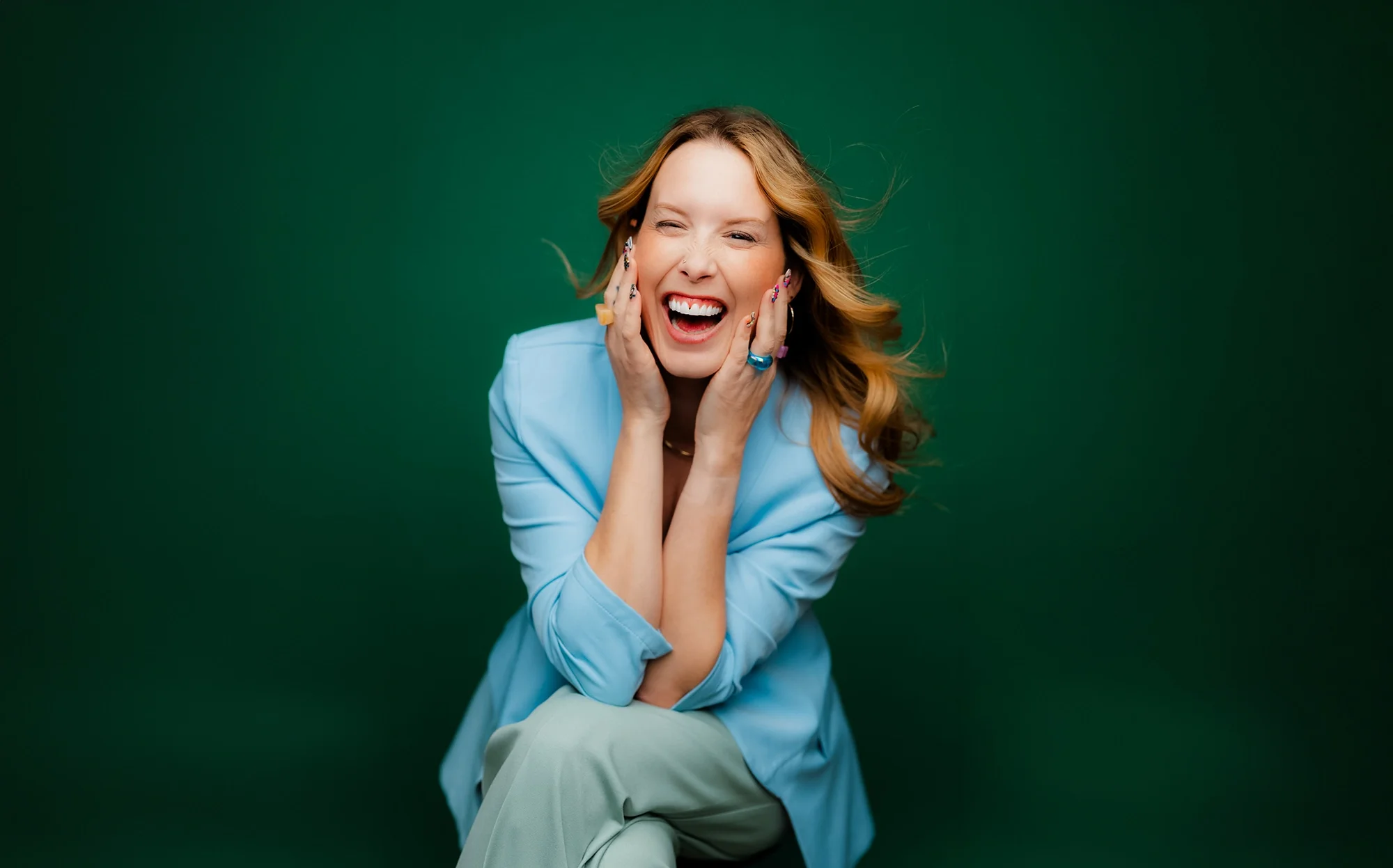 A woman with wavy red hair wearing a blue blazer and light-colored pants, laughing with her hands on her face against a green background.
