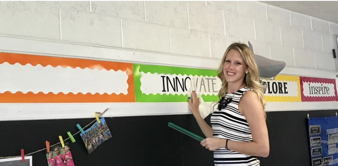 A woman with blonde hair in a black and white striped sleeveless top standing in a classroom, smiling, holding a green ruler, next to colorful academic banners on the wall that read 'Innovate,' 'Explore,' and 'Inspire.'