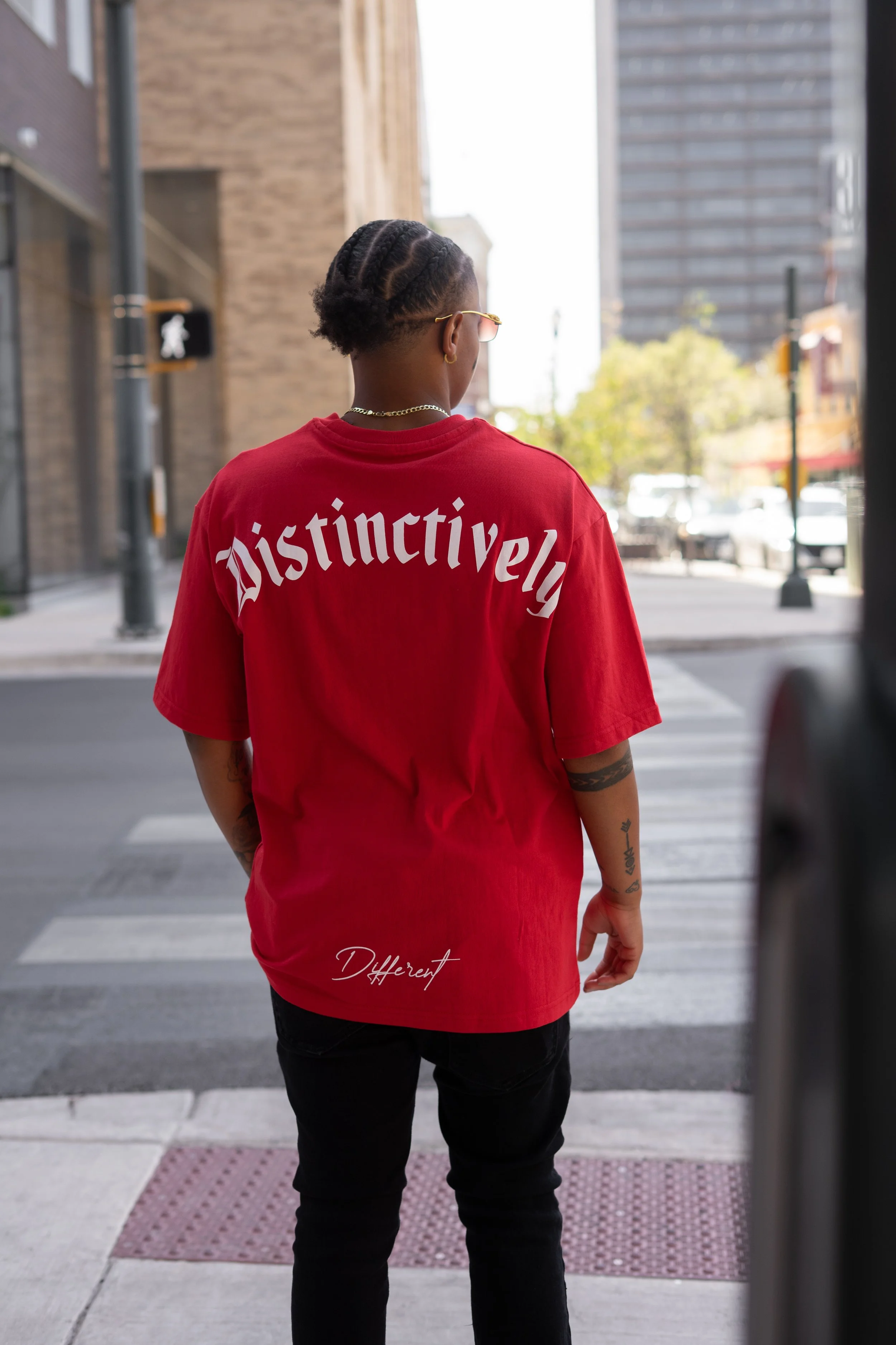 Person with braided hair wearing glasses and a red T-shirt with the word 'Distinctively' on the back and 'Different' at the bottom, standing at a city crosswalk.