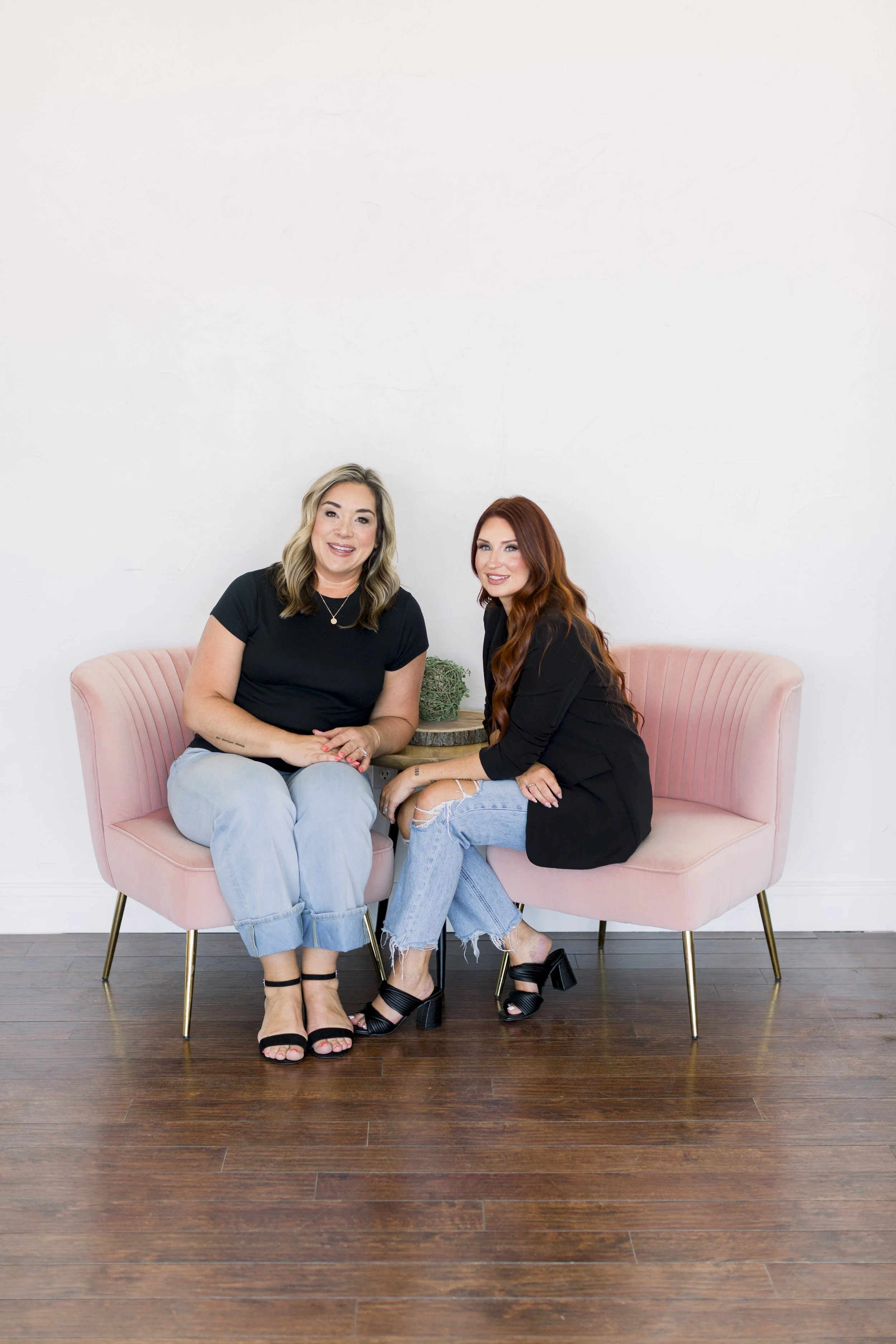 Two women sitting on a pink velvet loveseat with a small round wooden table between them, against a plain white wall, smiling at the camera.