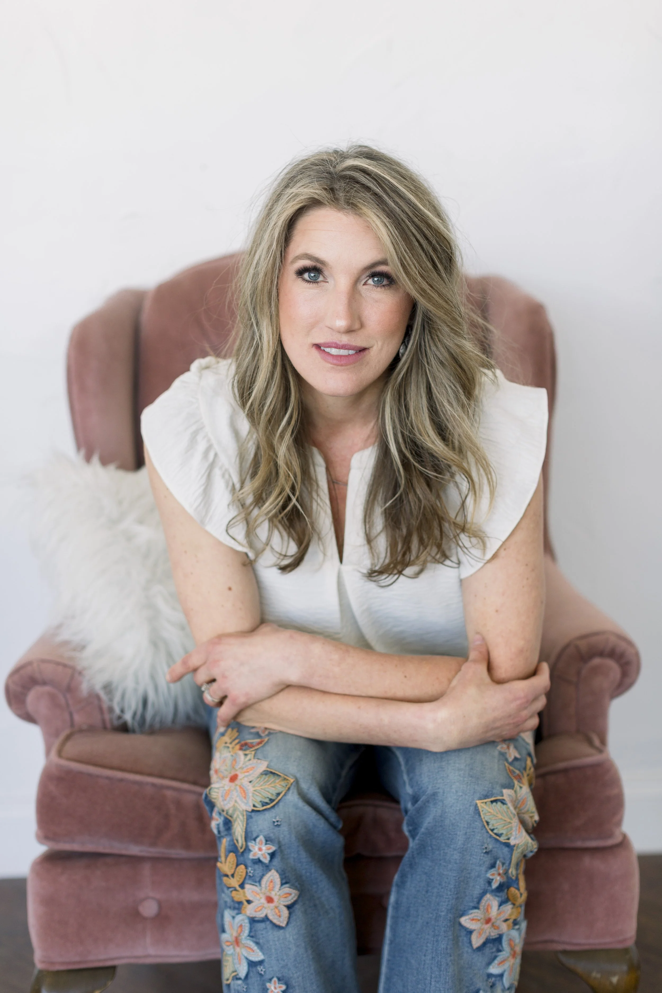 A woman with blonde hair, blue eyes, and a white top sitting on a pink velvet armchair with a white furry pillow in a bright room.