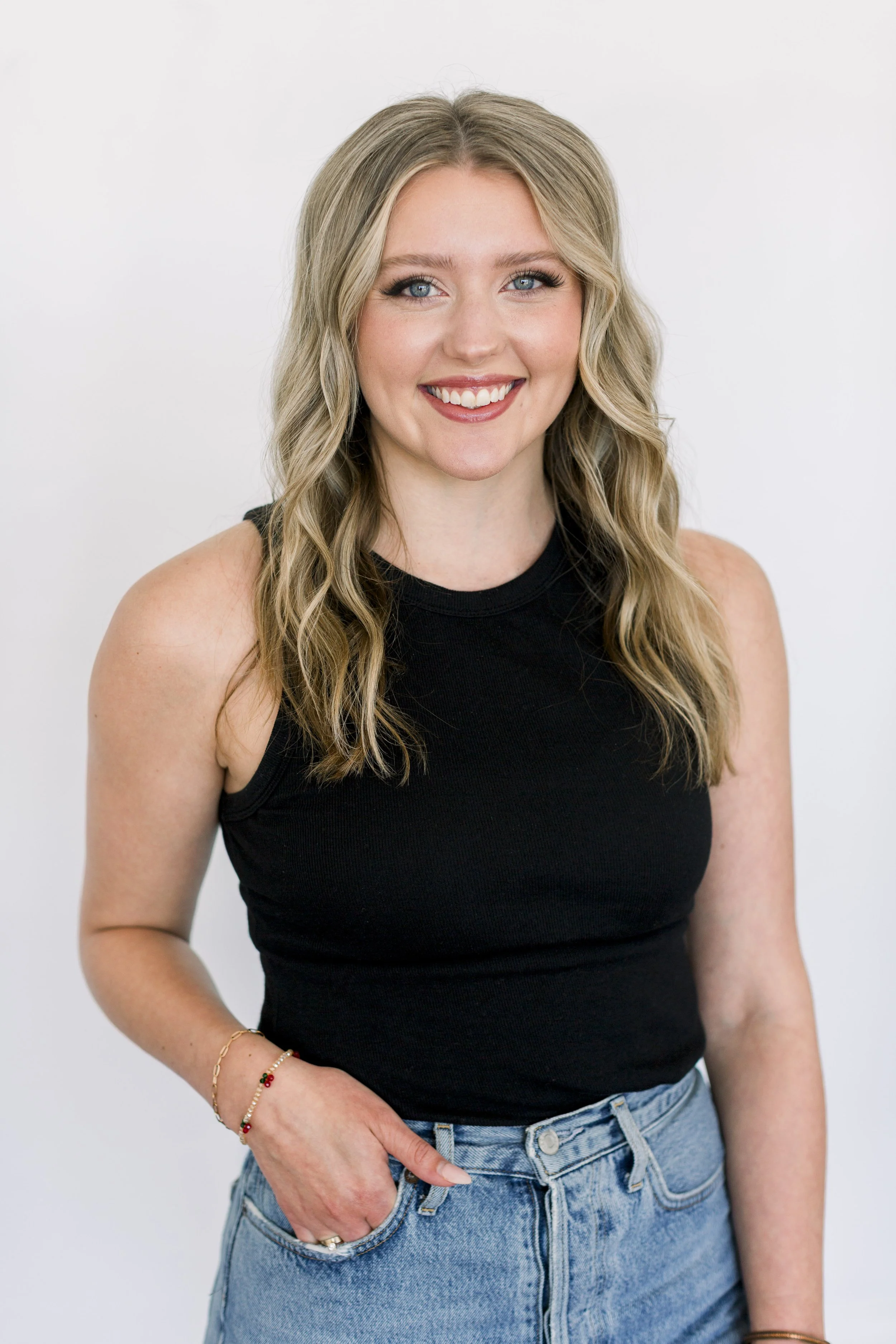 A young woman with long, wavy blonde hair, blue eyes, and fair skin, wearing a sleeveless black top and blue jeans. She is smiling and has one hand in her pocket.
