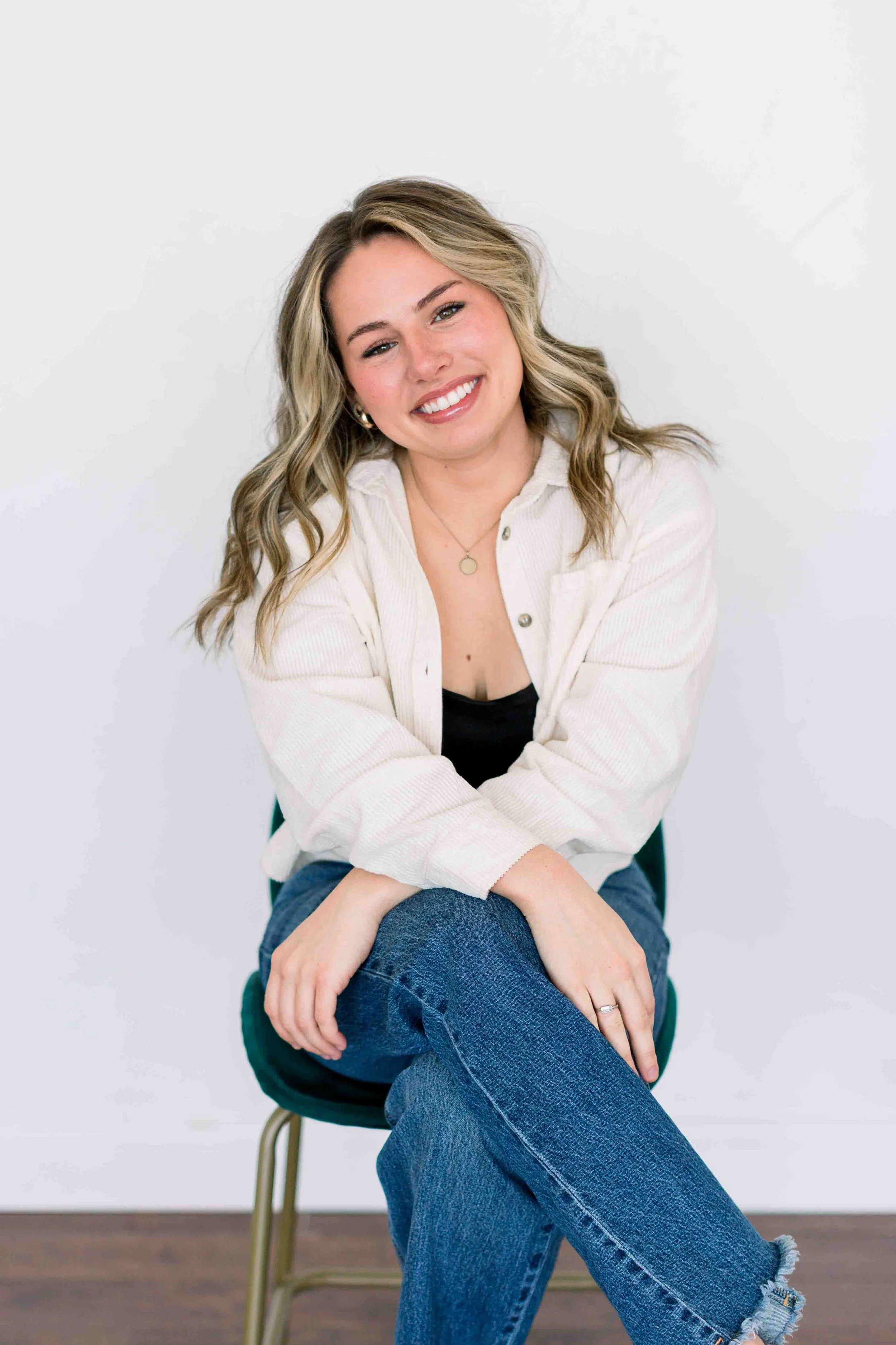 A young woman with blonde, wavy hair, smiling, sitting on a green chair against a plain white background, wearing a white button-up shirt, black top, and blue jeans.