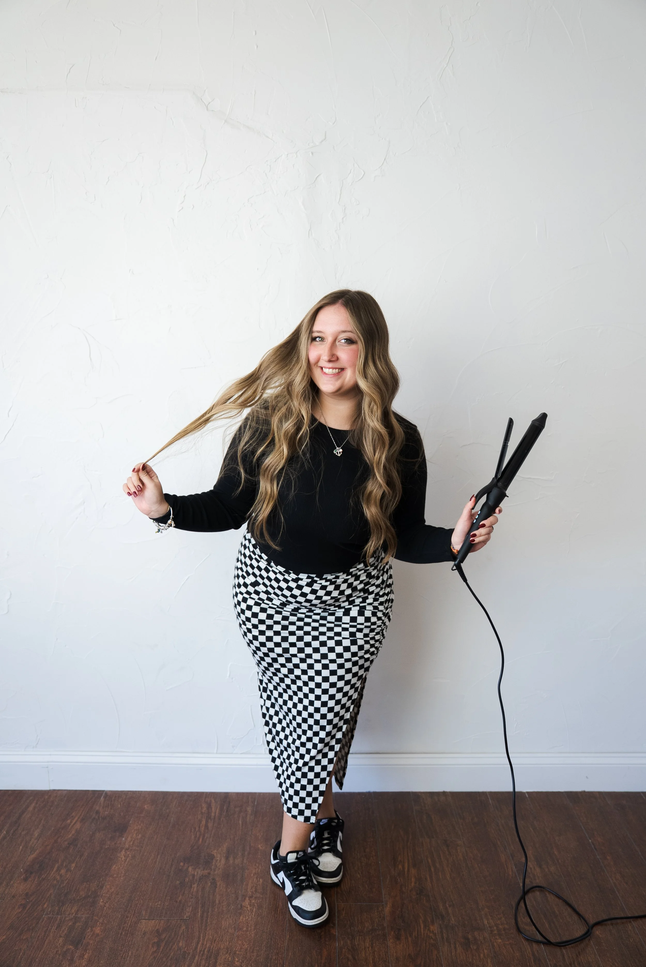 A young woman with long blonde hair, wearing a black long-sleeve top and black-and-white checkered skirt, is smiling while holding a curling iron in one hand and a section of her hair in the other. She is standing on a wooden floor against a plain white textured wall.