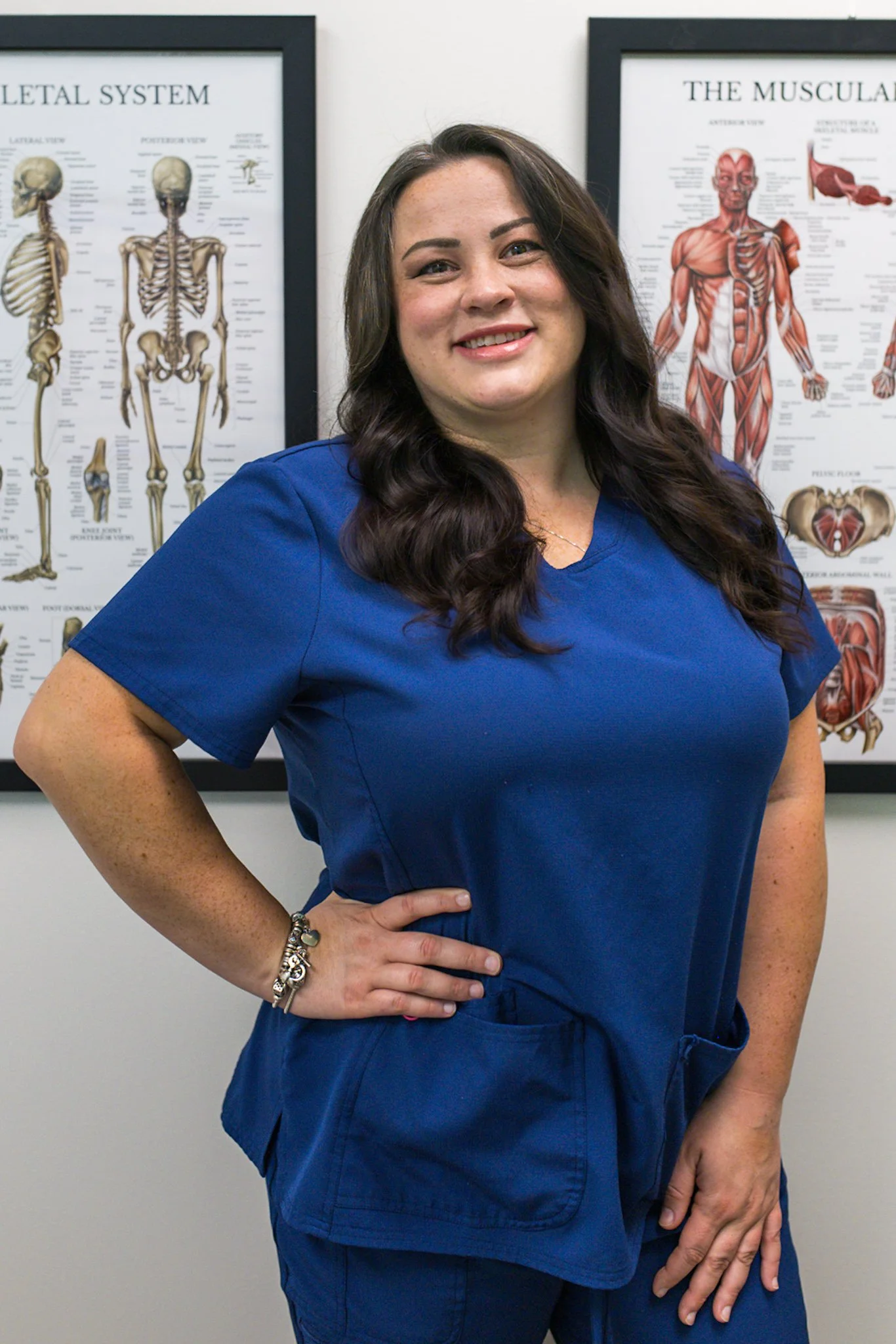 A woman in blue medical scrubs standing in front of anatomical posters of the skeletal and muscular systems.