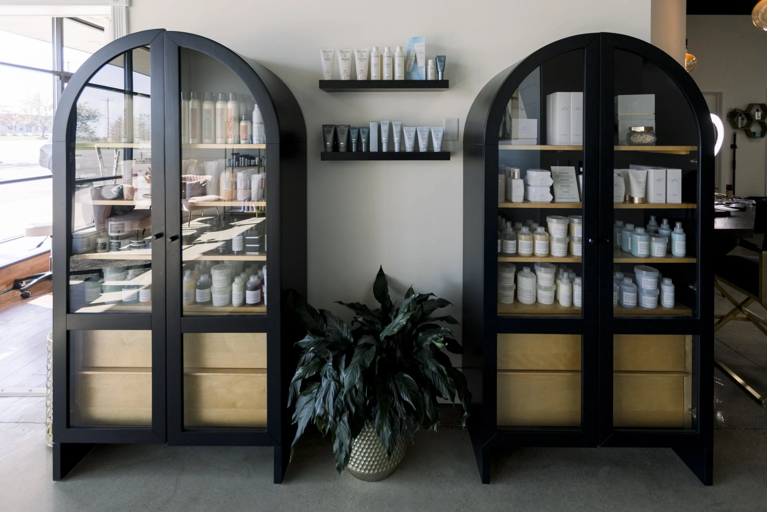 Two black display cabinets with glass doors, filled with skincare products, positioned on either side of a potted plant, with shelves of skincare items on the wall behind.