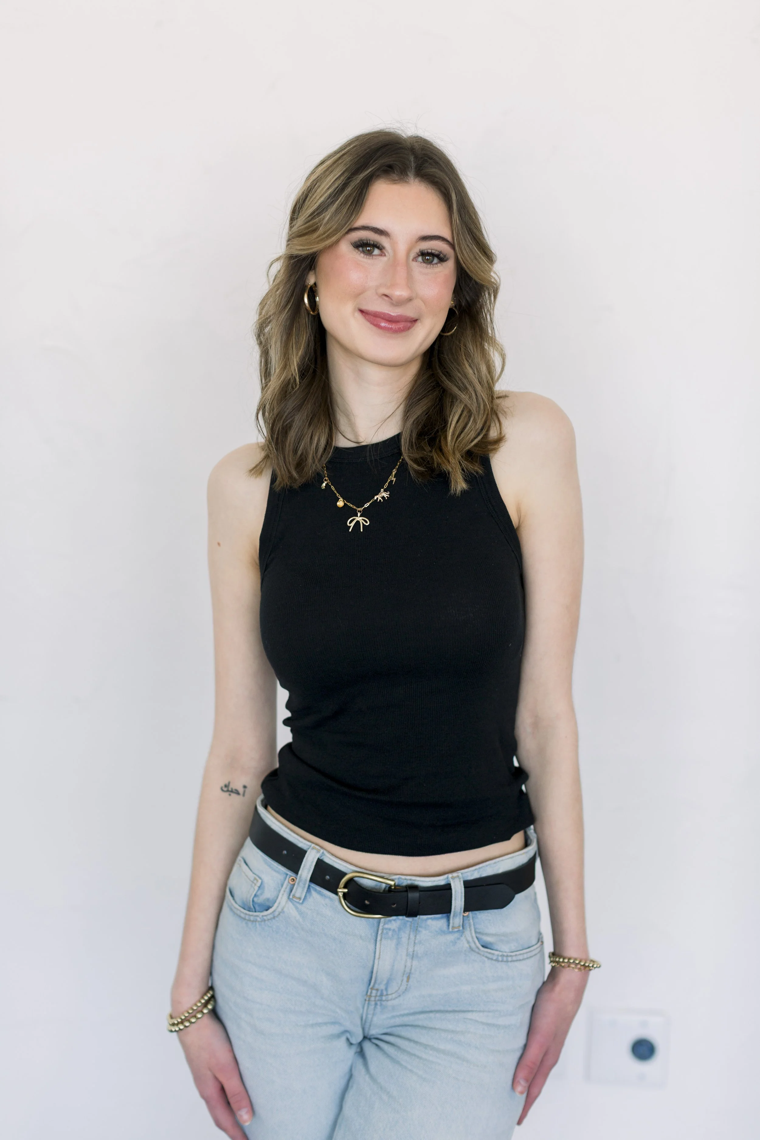 A young woman with shoulder-length wavy brown hair, wearing a sleeveless black top, light blue jeans, and gold jewelry, standing against a plain white background.