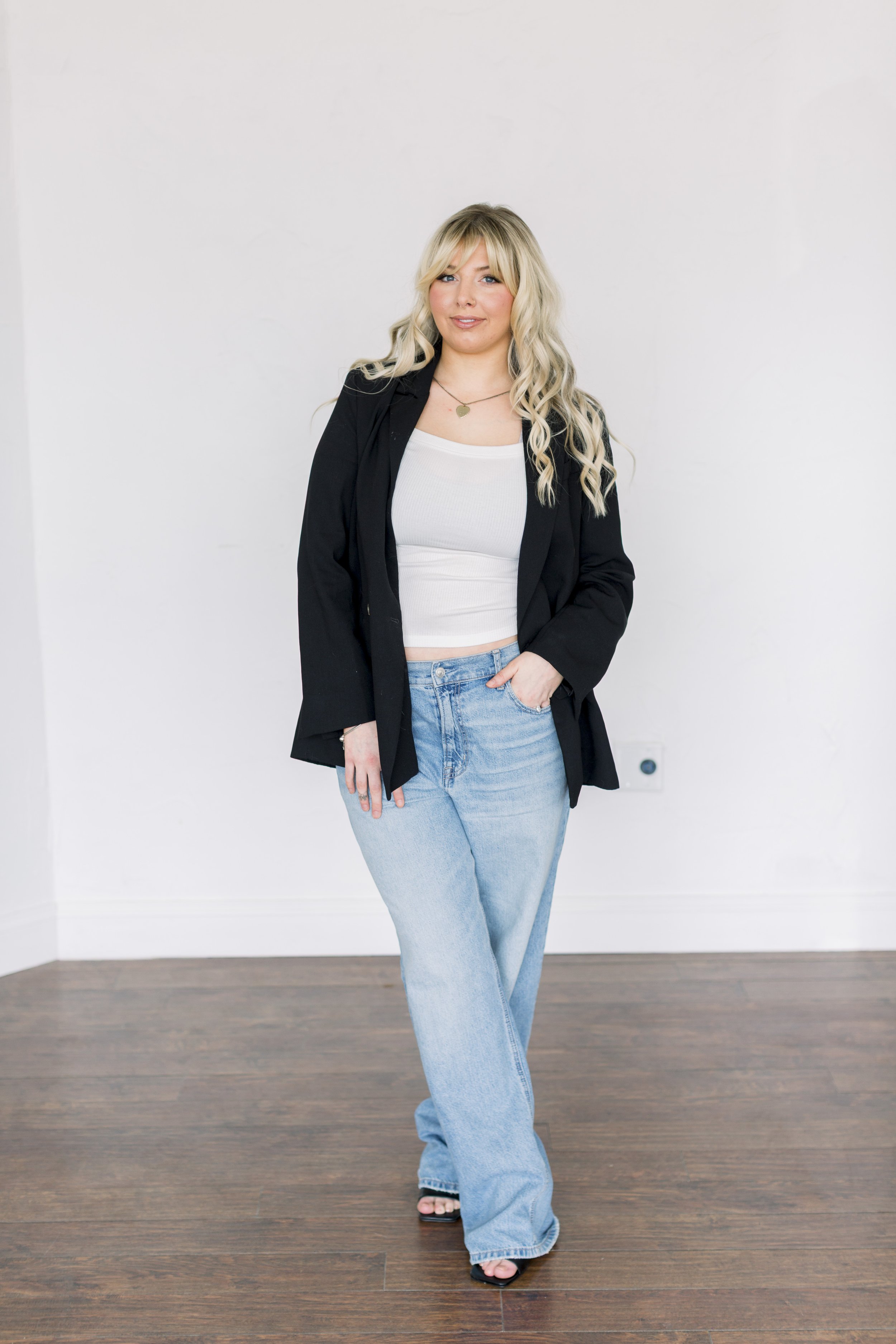 A woman with long blonde hair wearing a black blazer, white top, and light blue jeans standing on wooden floor against a plain white wall.
